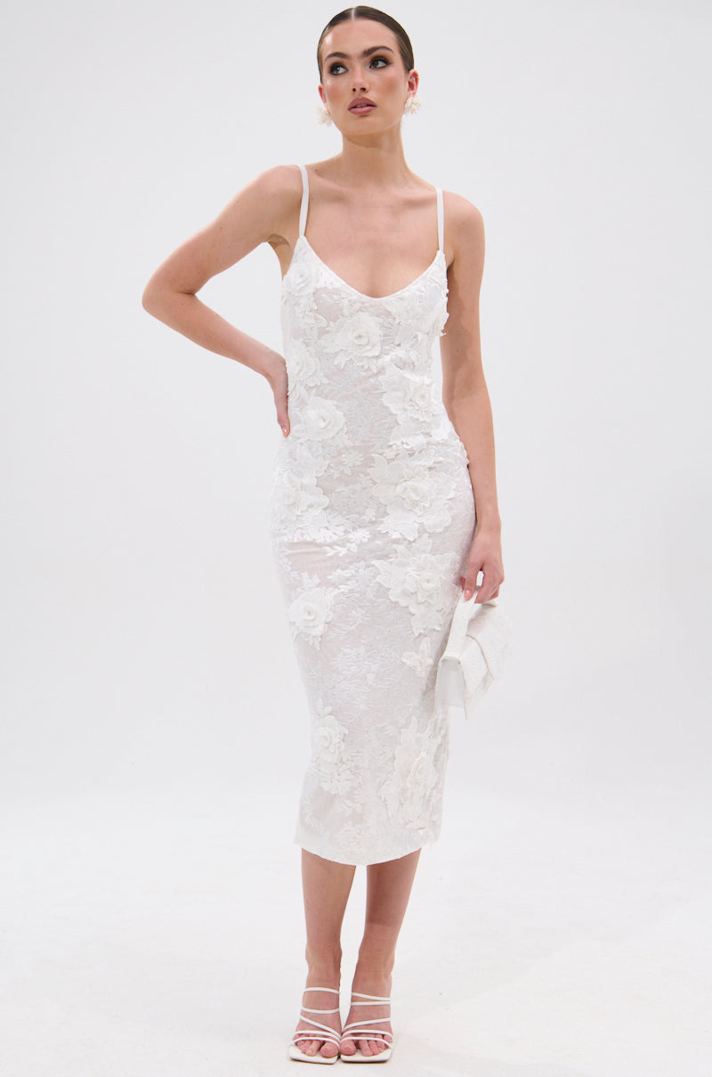 A woman stands against a white background wearing the IRIS FLORAL MIDI DRESS with 3D floral appliqués and thin straps. She pairs its bodycon silhouette with strappy white heels, carries a clutch, and looks confidently ahead. 7
