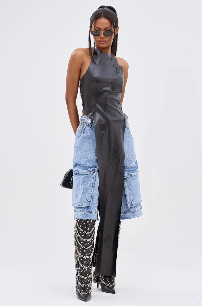 A woman wears the ADDIE FAUX LEATHER BLOUSE, a sleeveless black halter top with a long leather panel and cross appliqués, paired with blue denim cargo shorts, black knee-high boots with silver chains, sunglasses, and a black purse.