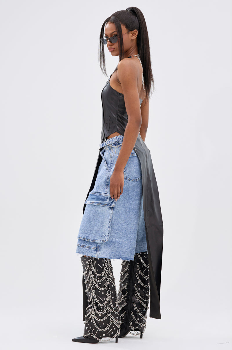 A woman with straight long hair wears the ADDIE FAUX LEATHER BLOUSE, light blue denim cargo shorts over jeweled, chain-embellished black pants, black pointed heels, and small sunglasses against a plain white background.