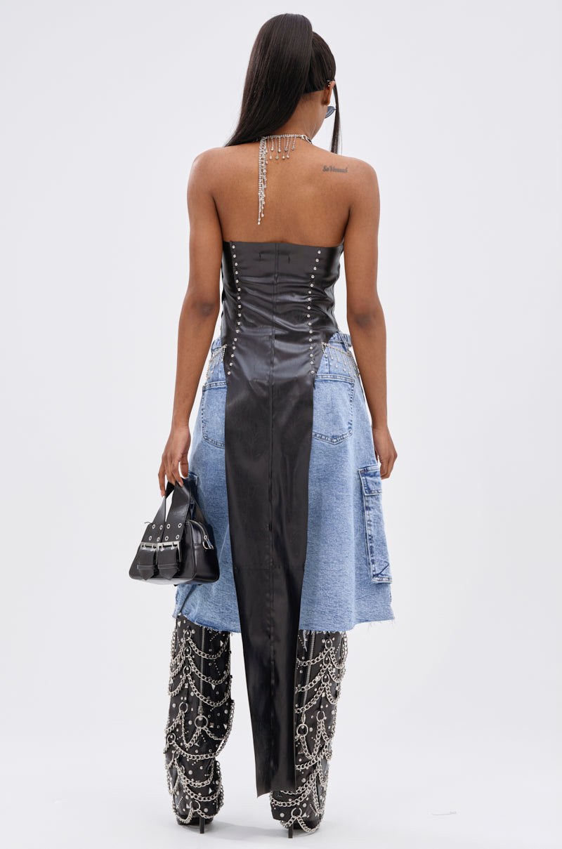 A woman faces away, wearing the ADDIE FAUX LEATHER BLOUSE with a halter neckline and long tail, paired with a knee-length denim skirt, embellished pants, sunglasses, and holding a small black handbag. Her hair is straight and long.
