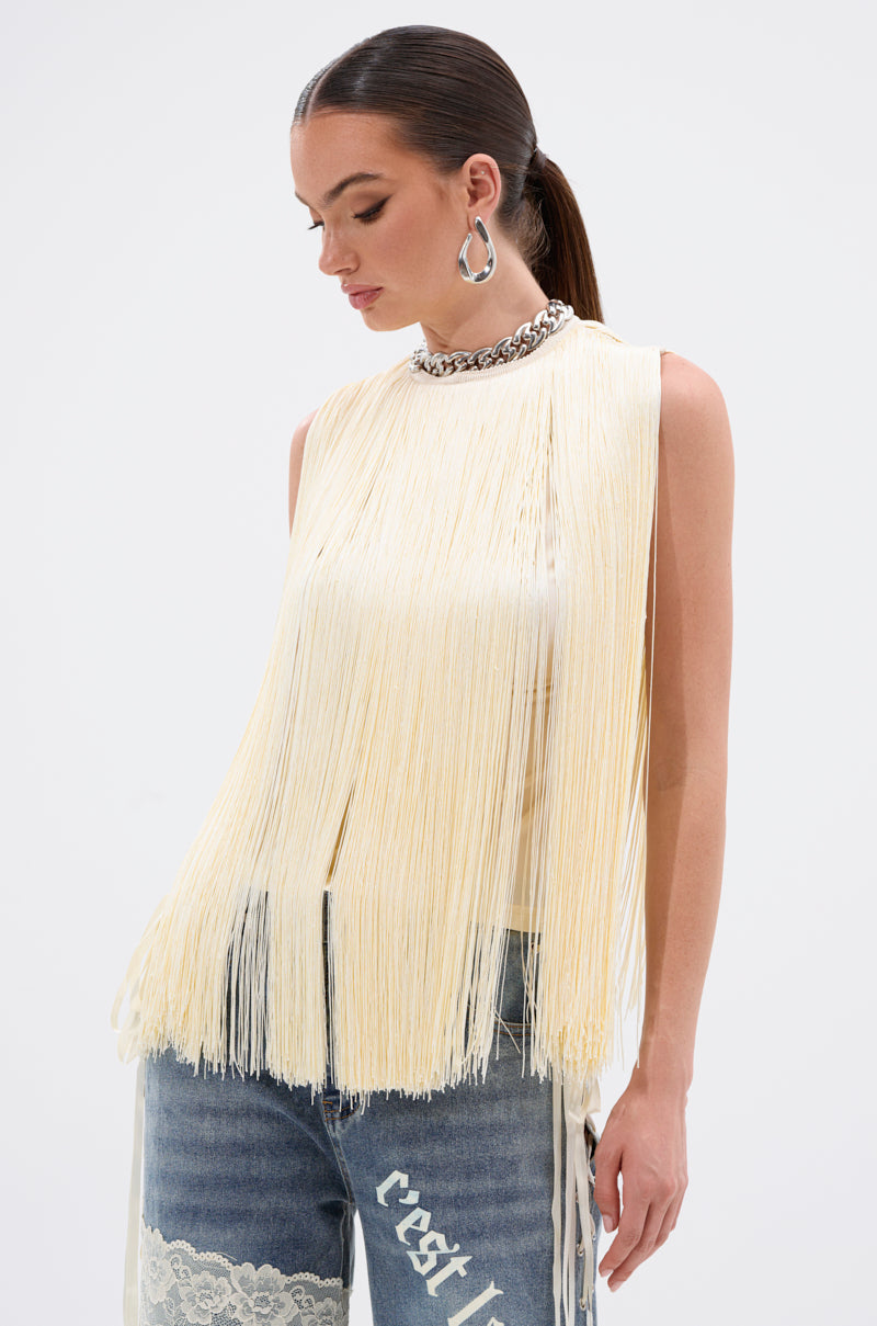 A woman wears the SUNSHINE LOVER FRINGE BLOUSE in butter yellow with chunky silver jewelry and lace-patched blue jeans, looking down against a white background—a summer staple.