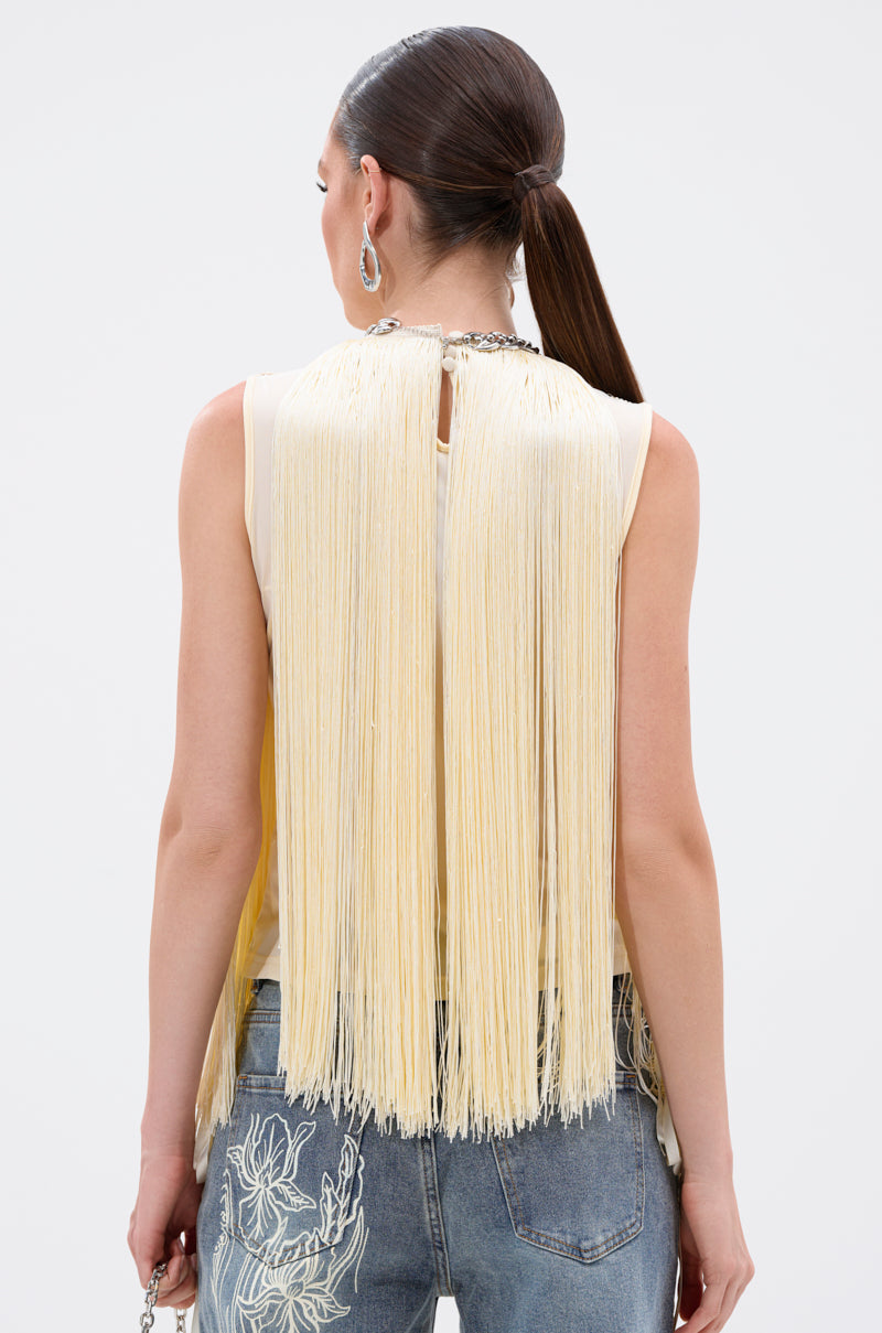A woman with long brown hair in a ponytail is seen from the back in the SUNSHINE LOVER FRINGE BLOUSE, a butter-yellow top, paired with blue jeans featuring white floral embroidery—summer style perfection.