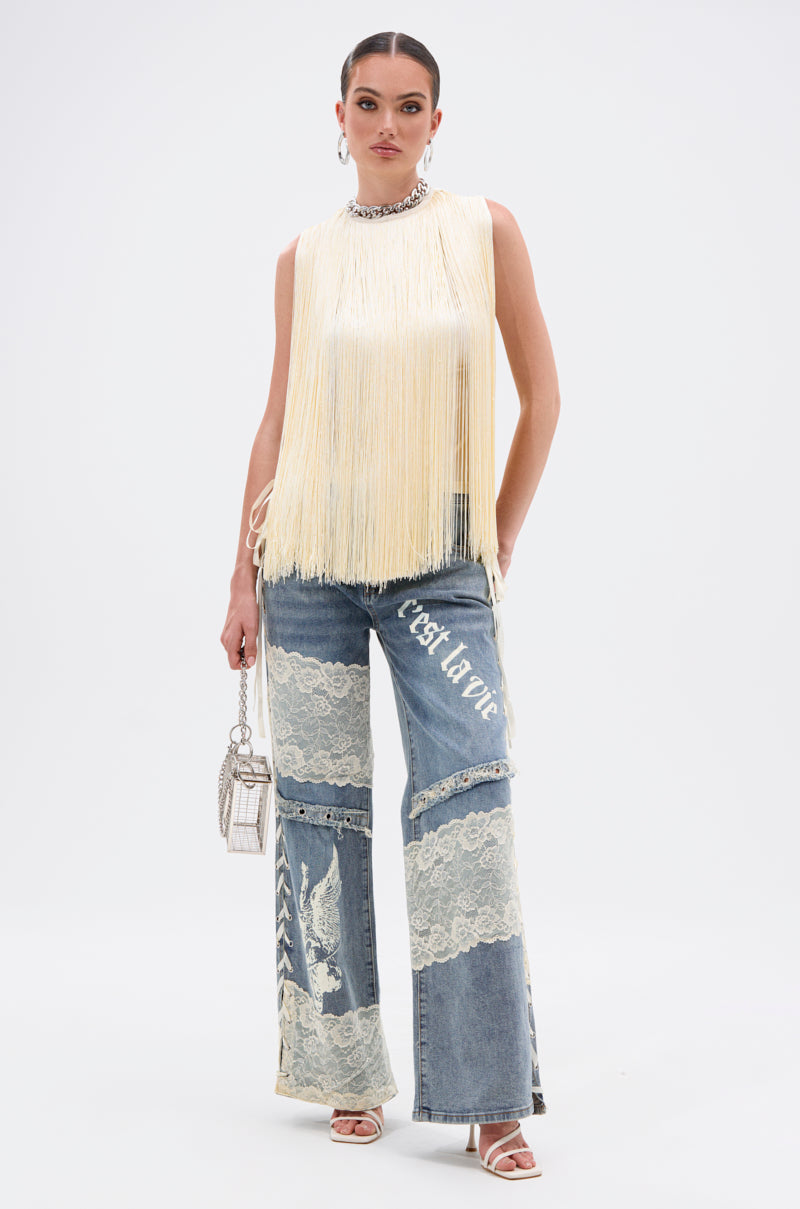 A woman stands against a white background wearing the SUNSHINE LOVER FRINGE BLOUSE in butter-yellow, styled with wide-leg jeans featuring lace panels and "c'est la vie," hoop earrings, heels, and a small purse.