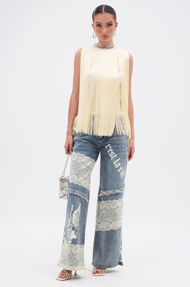 A woman stands against a plain background wearing the SUNSHINE LOVER FRINGE BLOUSE in butter-yellow, blue jeans with white lace panels and “C’est La Vie” text, white sandals, and holding a small metallic purse—perfect for summer.