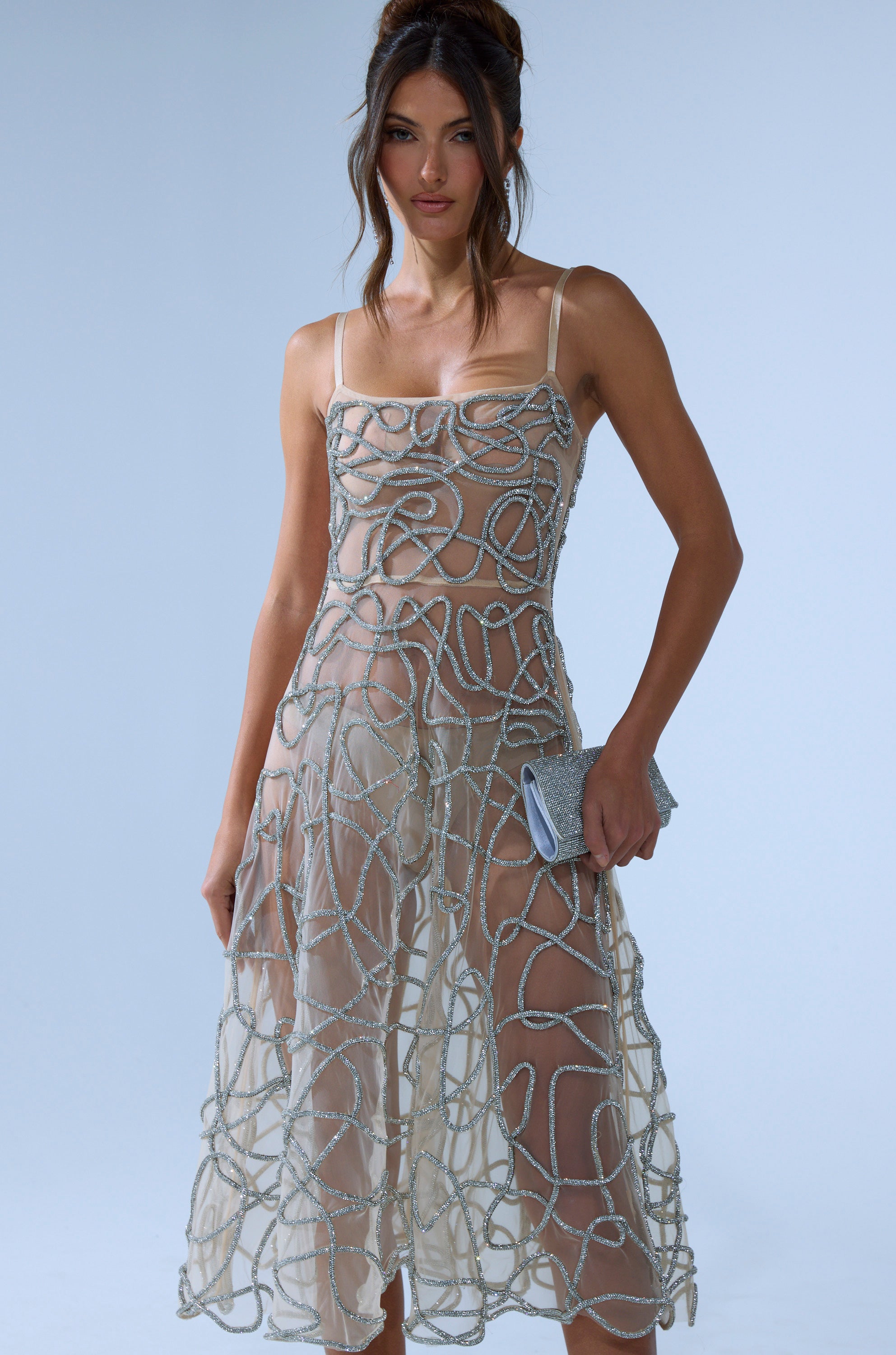 A woman wears the RIGHT THIS WAY RHINESTONE MIDI DRESS IN NUDE, featuring sheer mesh and silver rhinestone swirls, while holding a small silver clutch against a plain light blue background.
