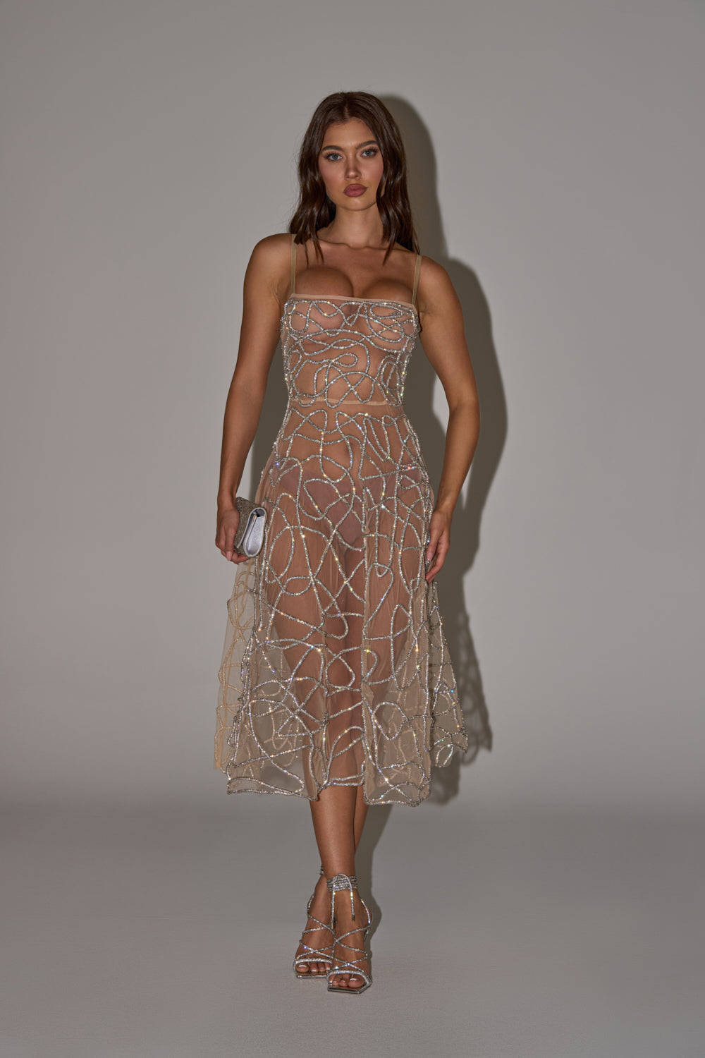 A woman stands against a plain backdrop in the RIGHT THIS WAY RHINESTONE MIDI DRESS IN NUDE, featuring sheer mesh with silver rhinestone swirls. She pairs it with strappy silver heels, a small clutch, and loose waves for an elegant evening look. 1