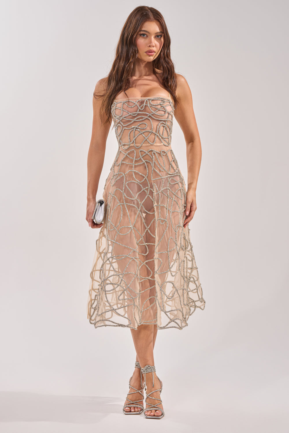 A woman wears the RIGHT THIS WAY RHINESTONE MIDI DRESS IN NUDE, a sheer mesh midi dress with rhinestone and silver swirling details, paired with strappy silver heels and a small silver clutch. 3
