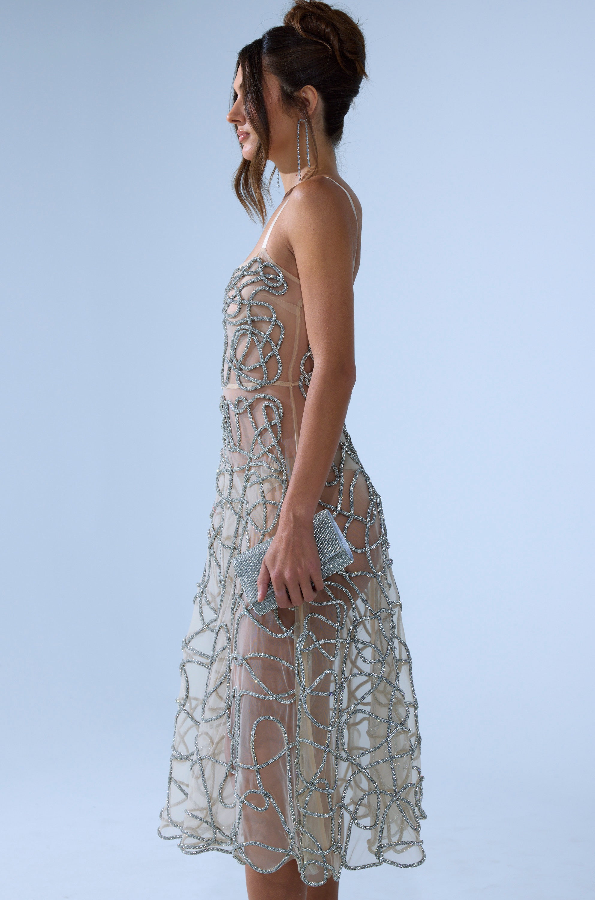 A woman in the RIGHT THIS WAY RHINESTONE MIDI DRESS IN NUDE, featuring sheer mesh and silver beadwork, stands in profile with her hair up, long earrings, and a sparkly clutch against a plain light blue background.
