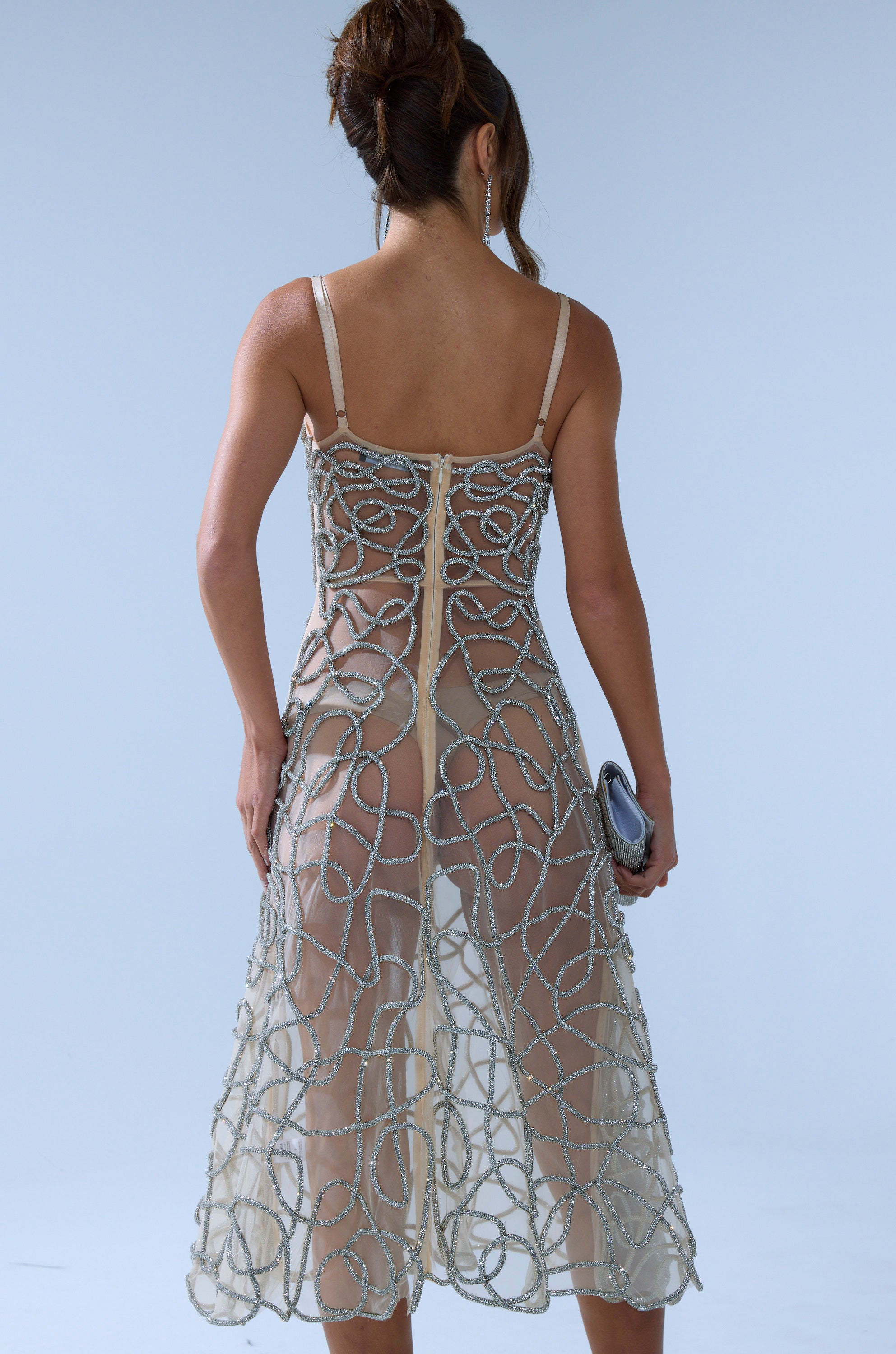 A woman with her hair in an updo wears the RIGHT THIS WAY RHINESTONE MIDI DRESS IN NUDE, a sleeveless sheer mesh dress with swirling silver beadwork, holding a small clutch and facing away from the camera against a light blue background.