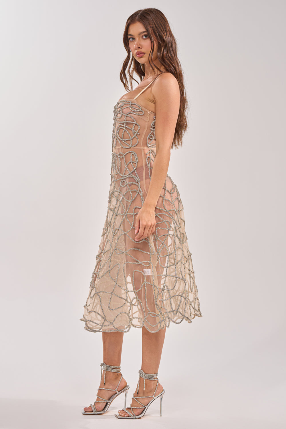 A woman with long, wavy brown hair and a neutral expression poses sideways against a plain background in the RIGHT THIS WAY RHINESTONE MIDI DRESS IN NUDE, featuring sheer mesh with silver embellishments and strappy high-heeled sandals. 5