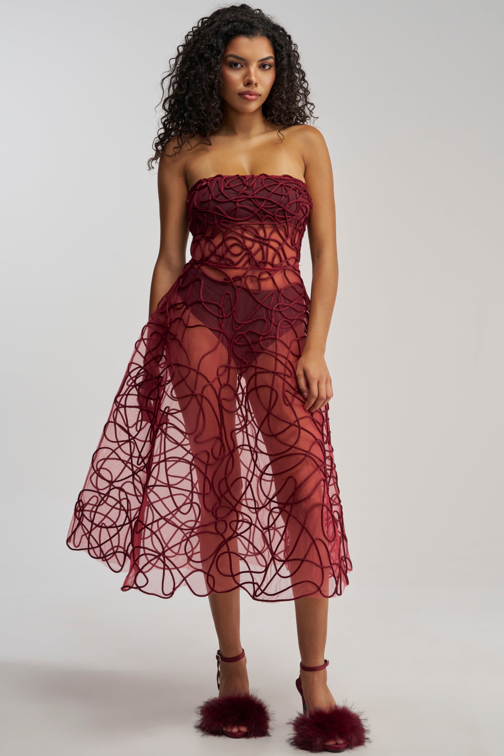 A woman stands against a plain light background wearing the MAYRA ABSTRACT MIDI DRESS IN BURGUNDY—an elegant burgundy mesh, strapless midi dress featuring swirling abstract corded embroidery—paired with matching burgundy heels with fluffy accents, making it the perfect choice for formal events.