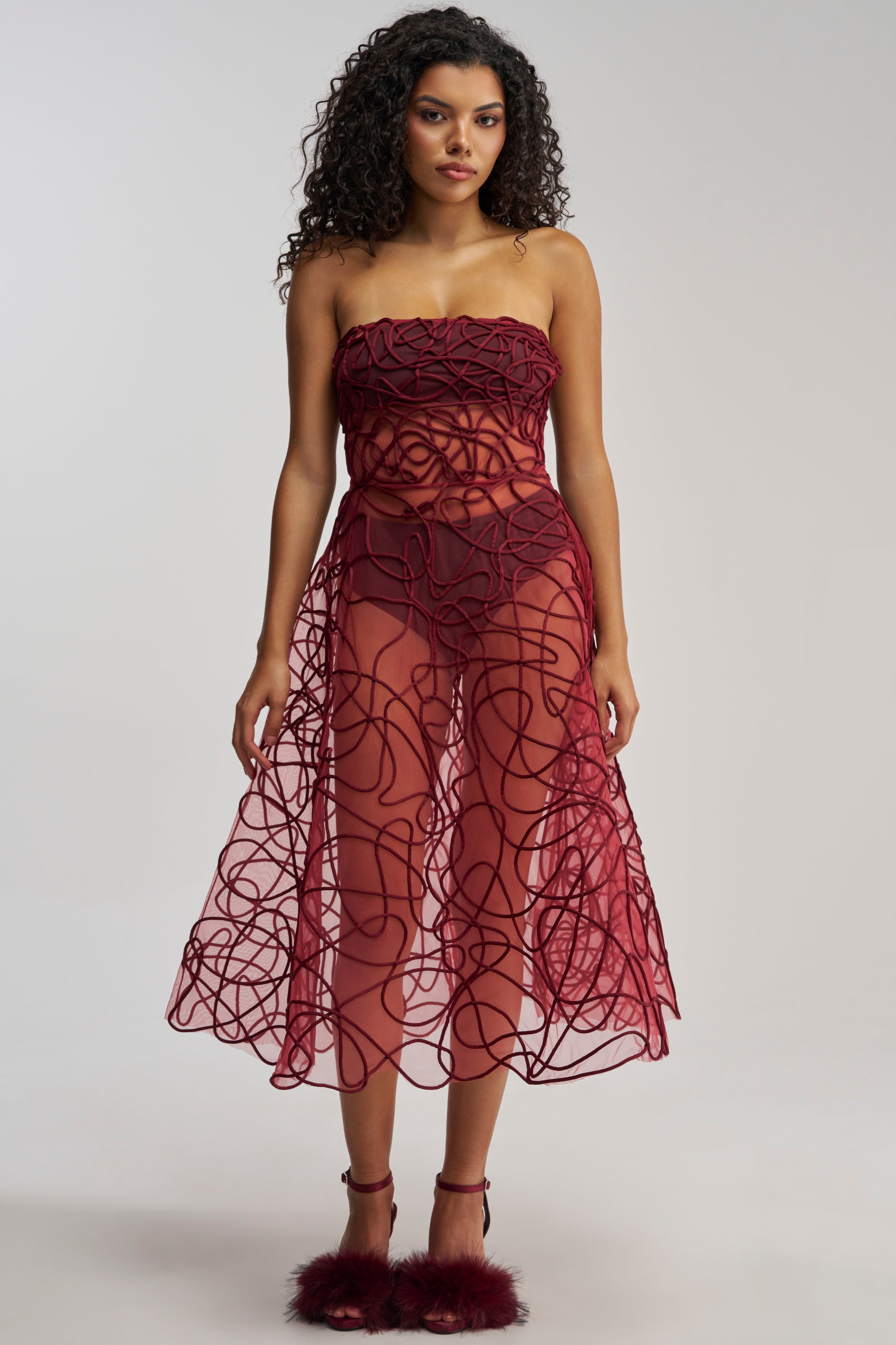 A woman with curly hair models the MAYRA ABSTRACT MIDI DRESS IN BURGUNDY, a strapless mesh midi dress showcasing abstract corded embroidery and a sheer skirt. She faces forward against a plain background, pairing the elegant dress with matching burgundy heels adorned with fluffy accents—an ideal choice for formal events.