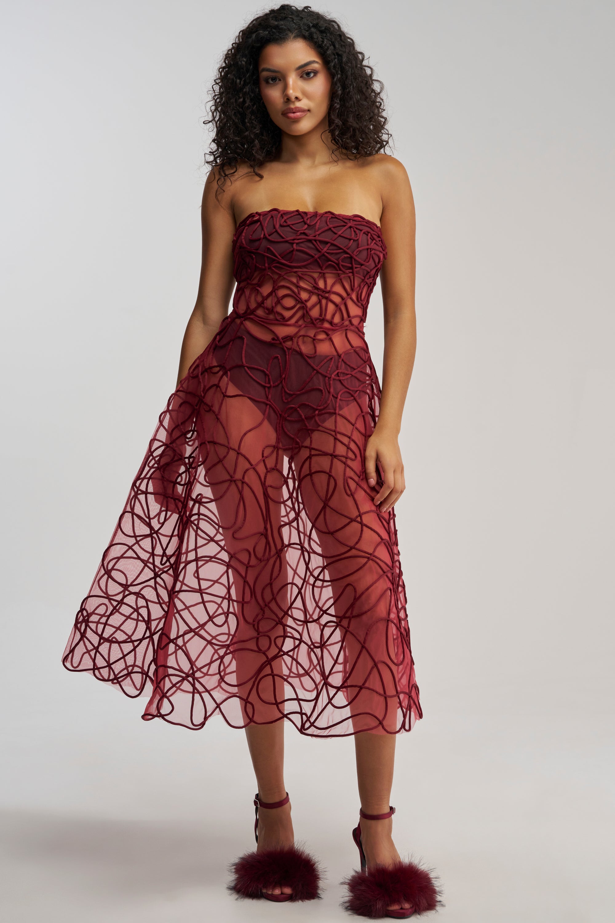 Wearing the MAYRA ABSTRACT MIDI DRESS IN BURGUNDY—featuring a strapless mesh design with swirling abstract patterns and delicate corded embroidery—a woman pairs it with matching burgundy feather-detailed heels as she stands against a plain light background.