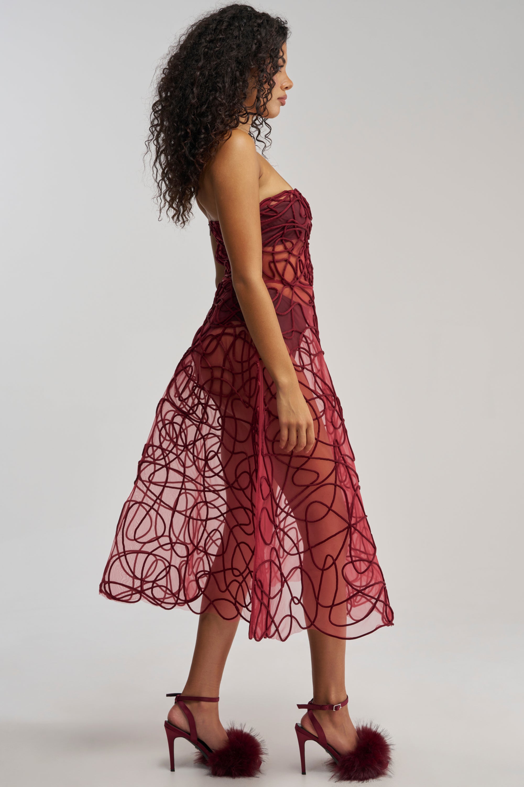 A woman with curly hair poses in profile, wearing the MAYRA ABSTRACT MIDI DRESS IN BURGUNDY—a sheer strapless midi dress featuring abstract swirling patterns—paired with matching high heels adorned with fluffy pom-poms, creating an effortlessly chic look for formal occasions.