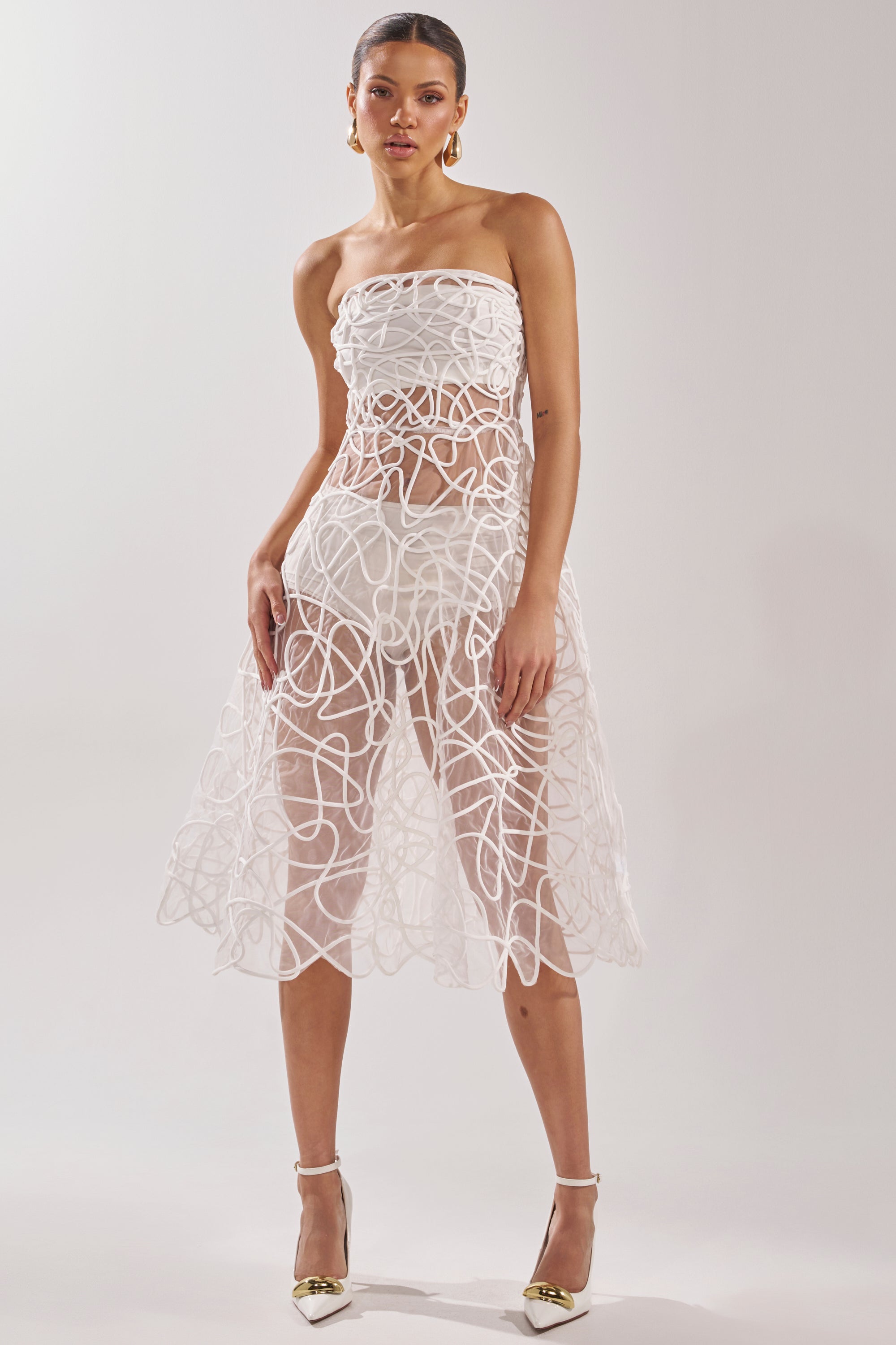 A woman poses against a plain background in the MAYA ABSTRACT MIDI DRESS IN WHITE, a strapless sheer midi dress with bold swirling white patterns. She completes the look with white high heels and gold hoop earrings. 8