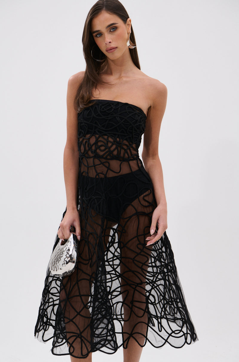 A woman stands against a plain white background wearing the MAYRA ABSTRACT MIDI DRESS— a strapless, sheer black mesh midi dress with abstract patterns, black undergarments, and holding a small silver handbag.