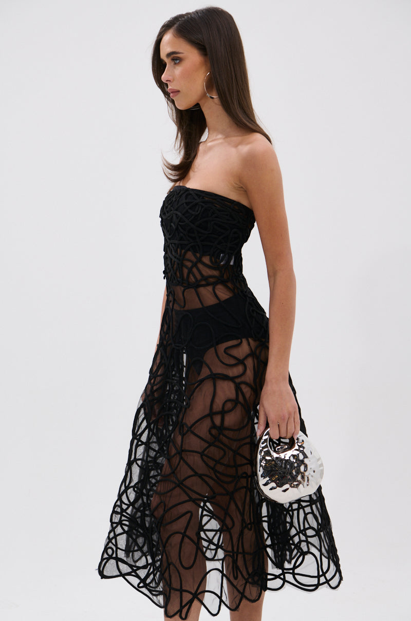 A woman wearing the MAYRA ABSTRACT MIDI DRESS, a strapless mesh midi dress with abstract swirling patterns, stands holding a small white and black handbag against a plain white background.
