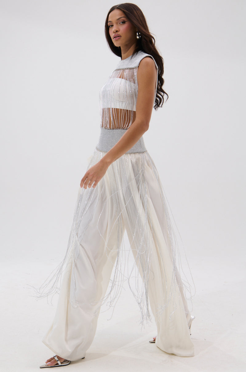 A woman confidently models the ARK SLEEVELESS CHAIN TOP— a white, sleeveless crop top with metallic fringe details—paired with matching wide-leg pants featuring sheer panels and long fringe against a plain white background. 1