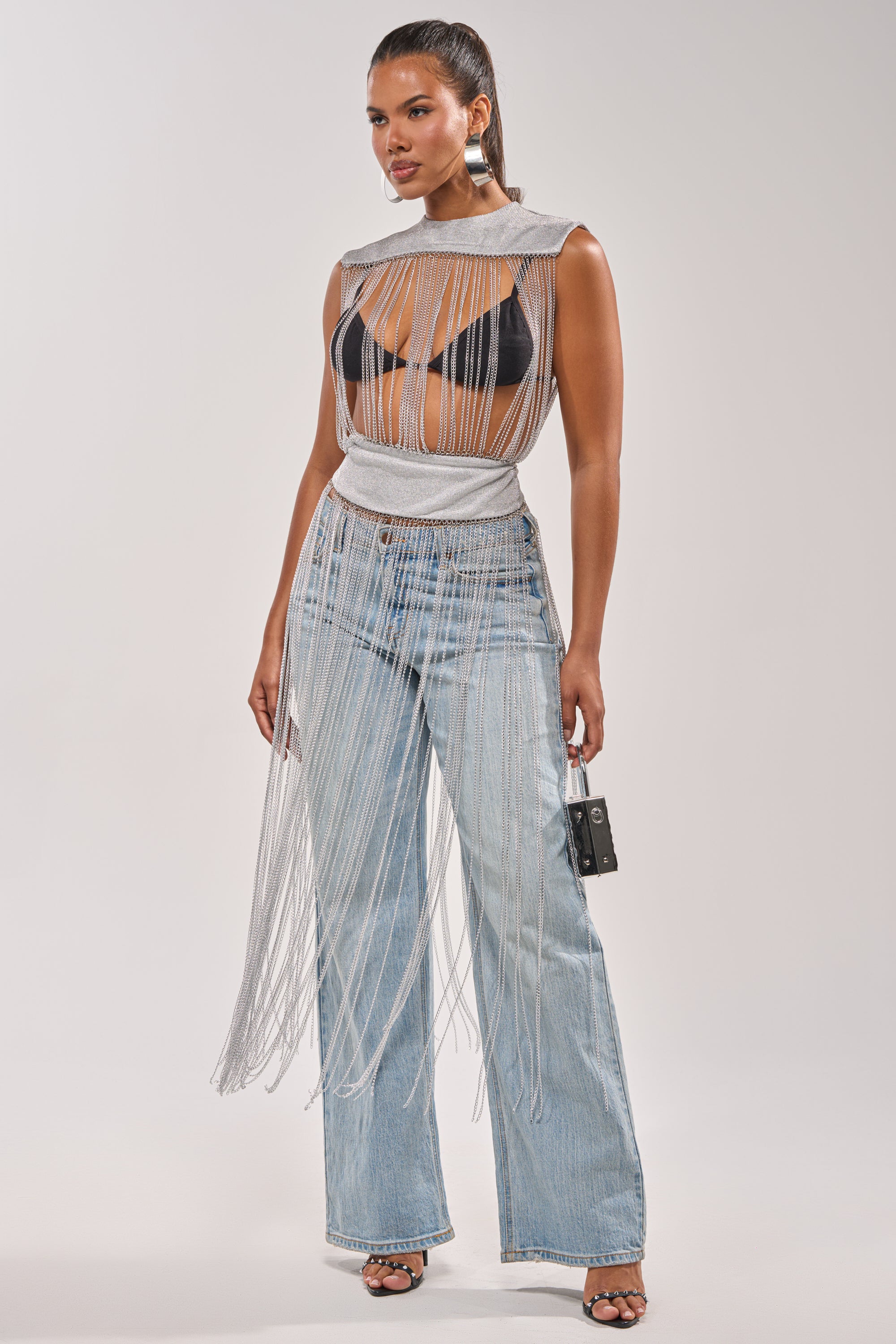 A woman models the ARK SLEEVELESS CHAIN TOP—a silver chain fringe, sleeveless statement top—paired with a black bralette, light blue high-waisted wide-leg jeans, black heeled sandals, a small black purse, and a ponytail. 0