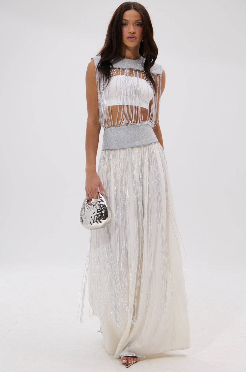 A woman wearing the ARK SLEEVELESS CHAIN TOP with metallic fringe details and holding a shiny silver clutch stands against a white background. She has long, dark hair and gazes forward with a relaxed expression. 2