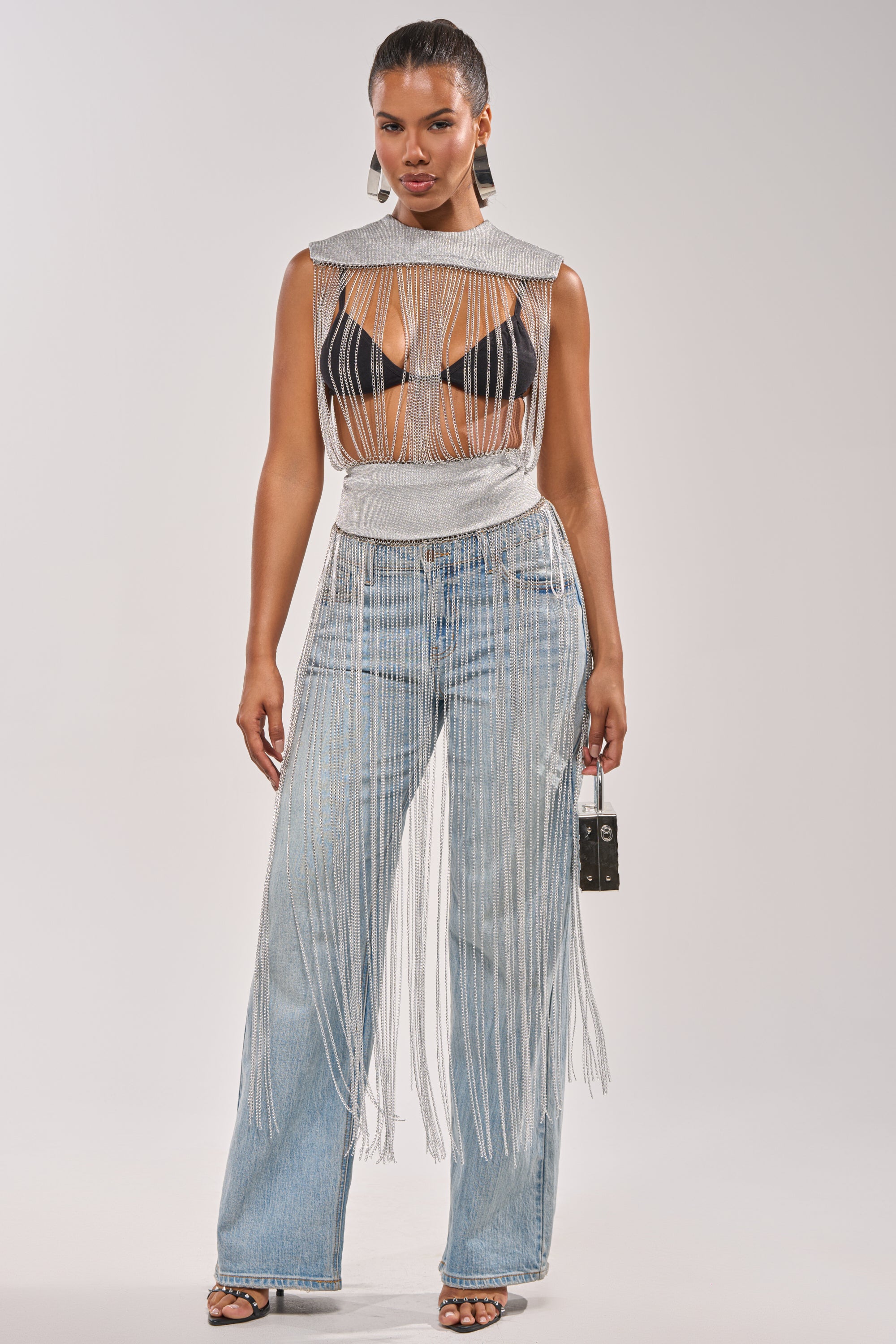 A woman exudes confidence in the ARK SLEEVELESS CHAIN TOP with silver fringe over a black bralette, styled with high-waisted, wide-leg jeans with matching fringe. She completes her concert-ready look with a small handbag and open-toe heels. 1