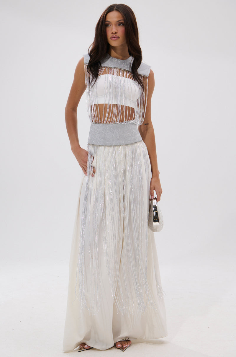 A woman poses elegantly against a white background in the ARK SLEEVELESS CHAIN TOP, styled with a long, flowing silver-fringed skirt, silver clutch, and heels. 3