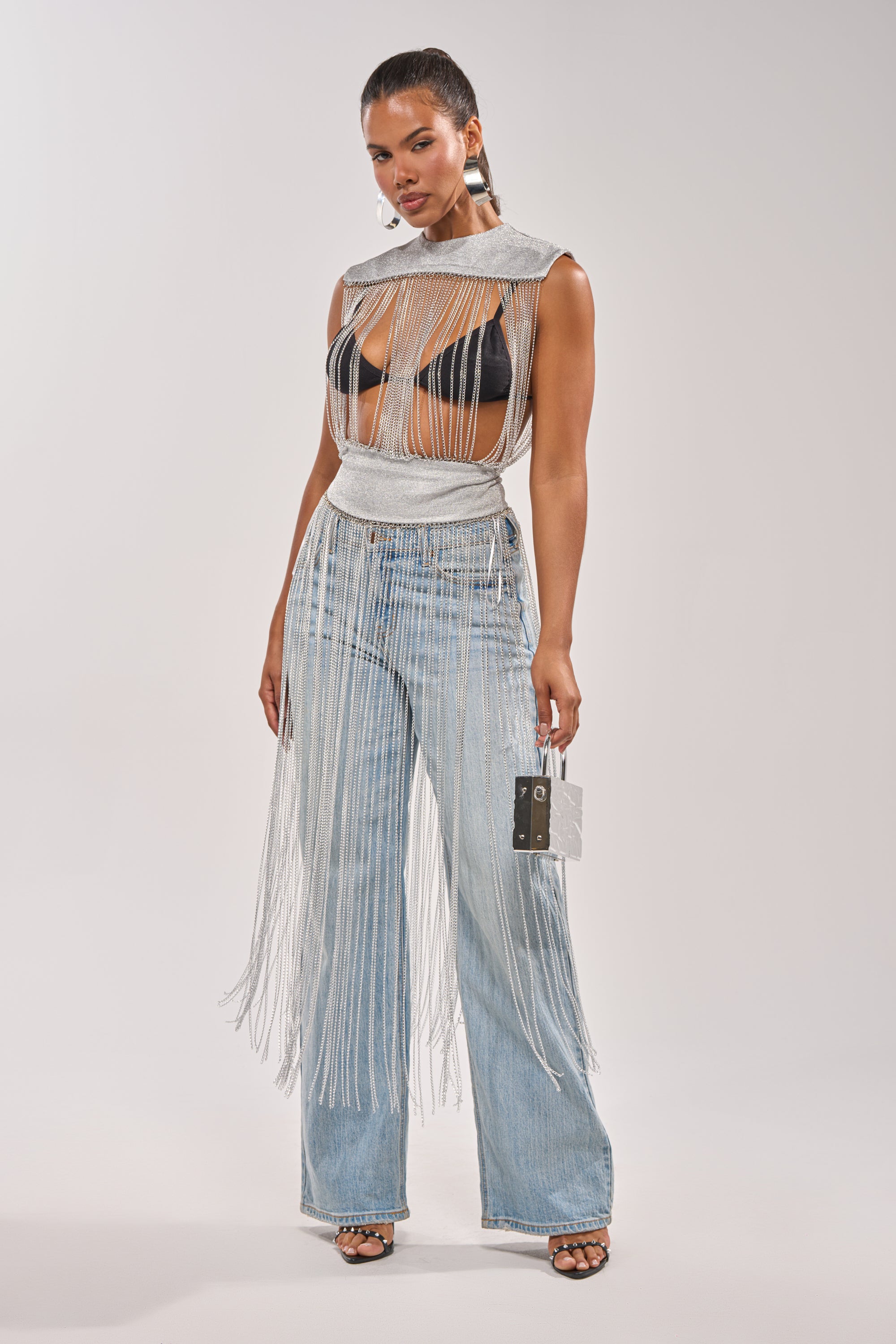 A woman exudes confidence in the ARK SLEEVELESS CHAIN TOP—a silver chain fringe top layered over a black bra, styled with high-waisted light blue fringe jeans, a small silver purse, and black heels against a plain background. 2