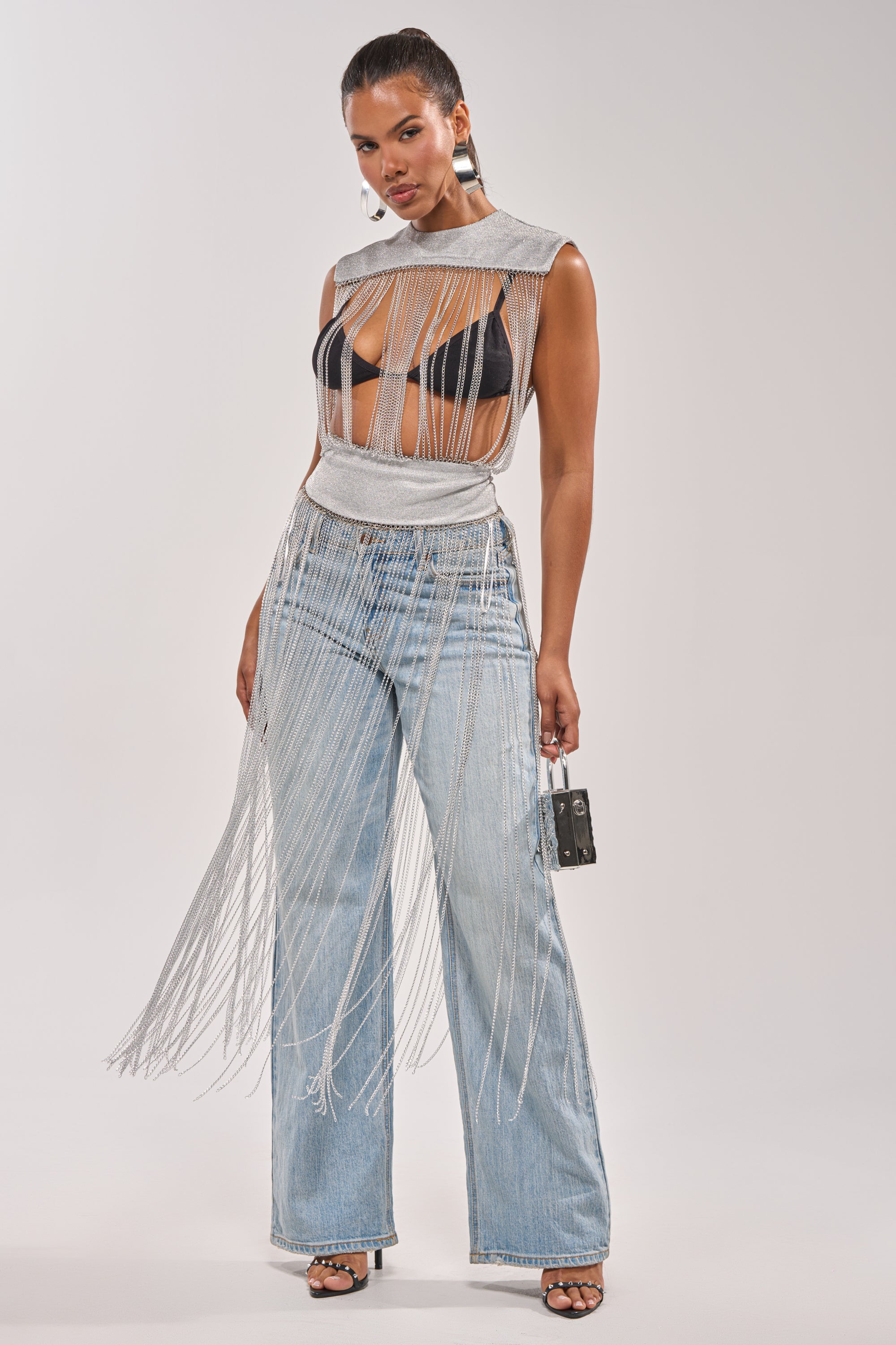 A woman models the ARK SLEEVELESS CHAIN TOP over a black bra with light blue wide-leg jeans and black heels, accessorized with a small black purse. Her hair is slicked back as she poses against a light gray background—ideal for bold concert looks. 3