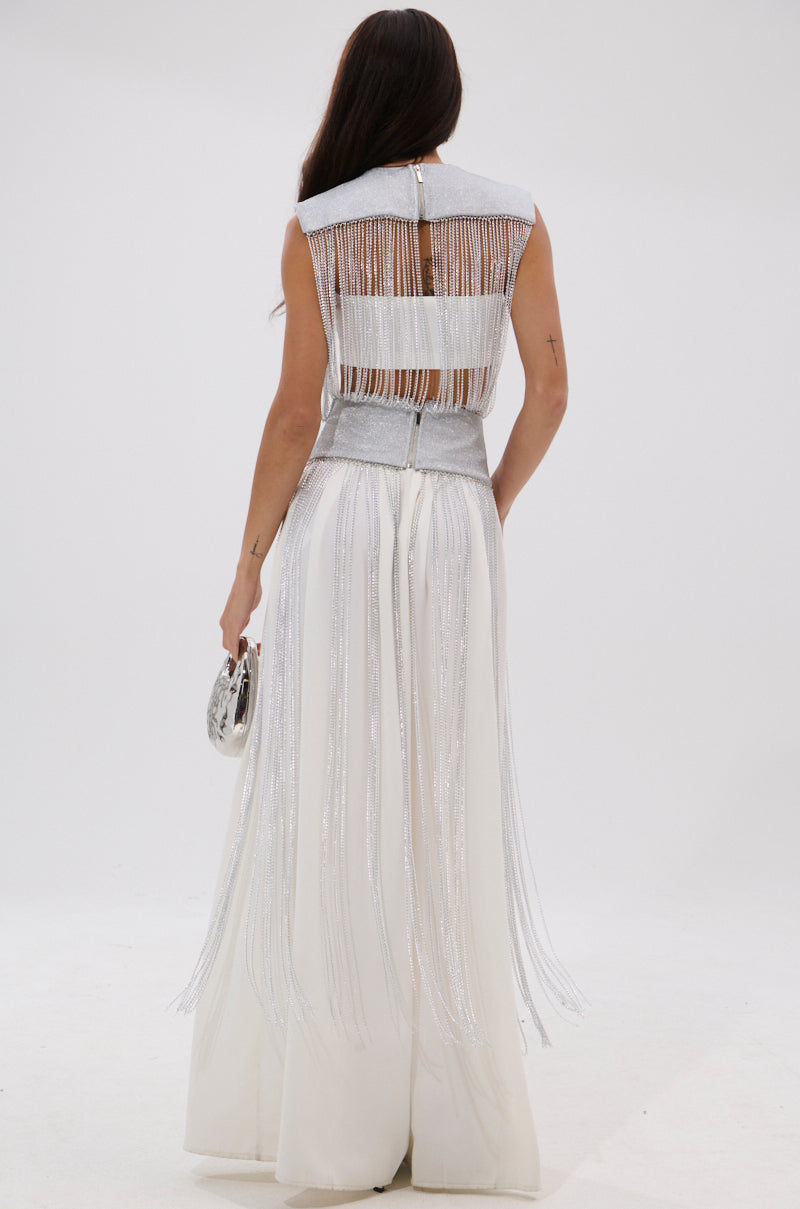 A woman with long dark hair stands facing away, wearing the ARK SLEEVELESS CHAIN TOP paired with a white fringed gown and holding a small silver clutch against a plain white background. 5