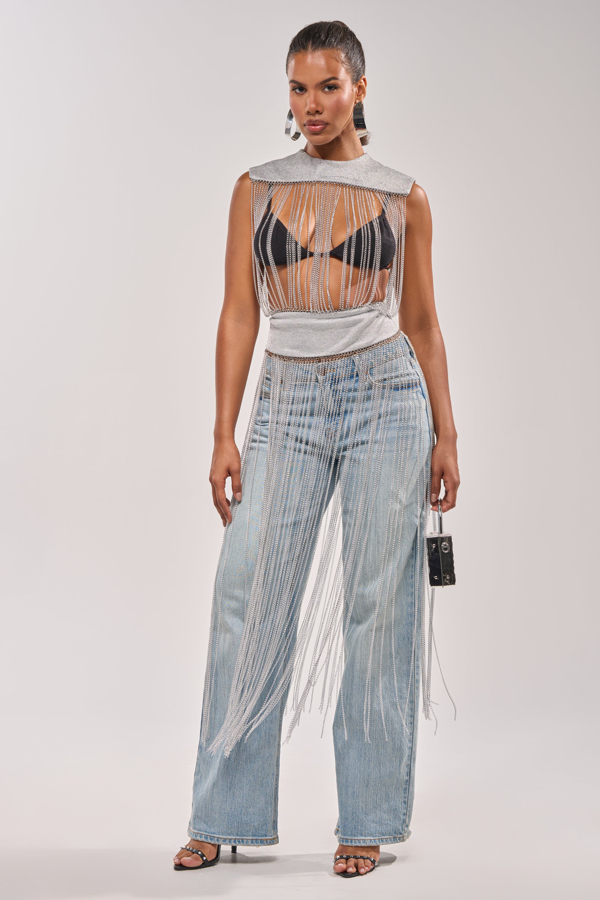 A woman exudes confidence in the ARK SLEEVELESS CHAIN TOP layered over a black bra, styled with high-waisted light blue wide-leg jeans and strappy heels. She completes her chic festival look with a sleek updo and a small handbag. 4