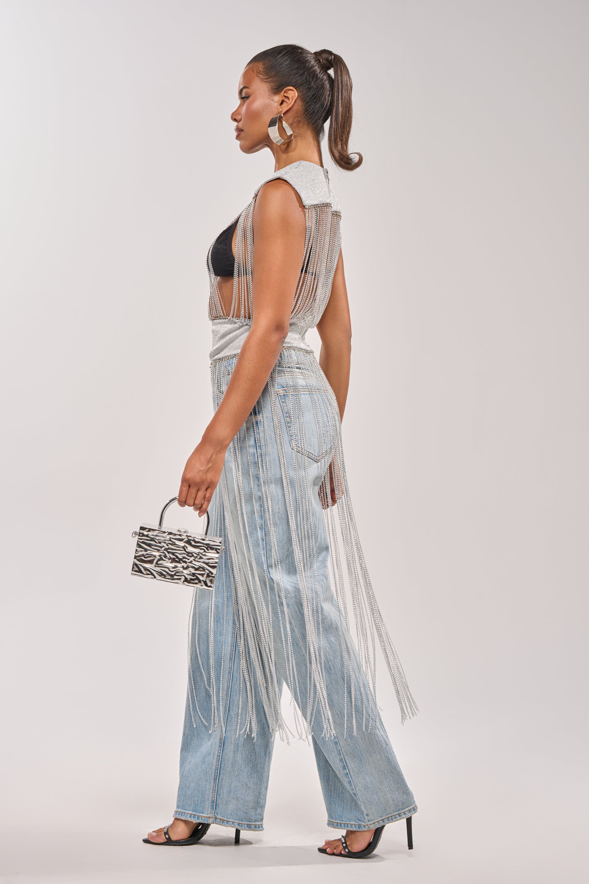 A woman stands in profile wearing the ARK SLEEVELESS CHAIN TOP over wide-leg jeans, high heels, and large hoop earrings, holding a small zebra-print handbag—an eye-catching concert look with her hair in a sleek ponytail. 5