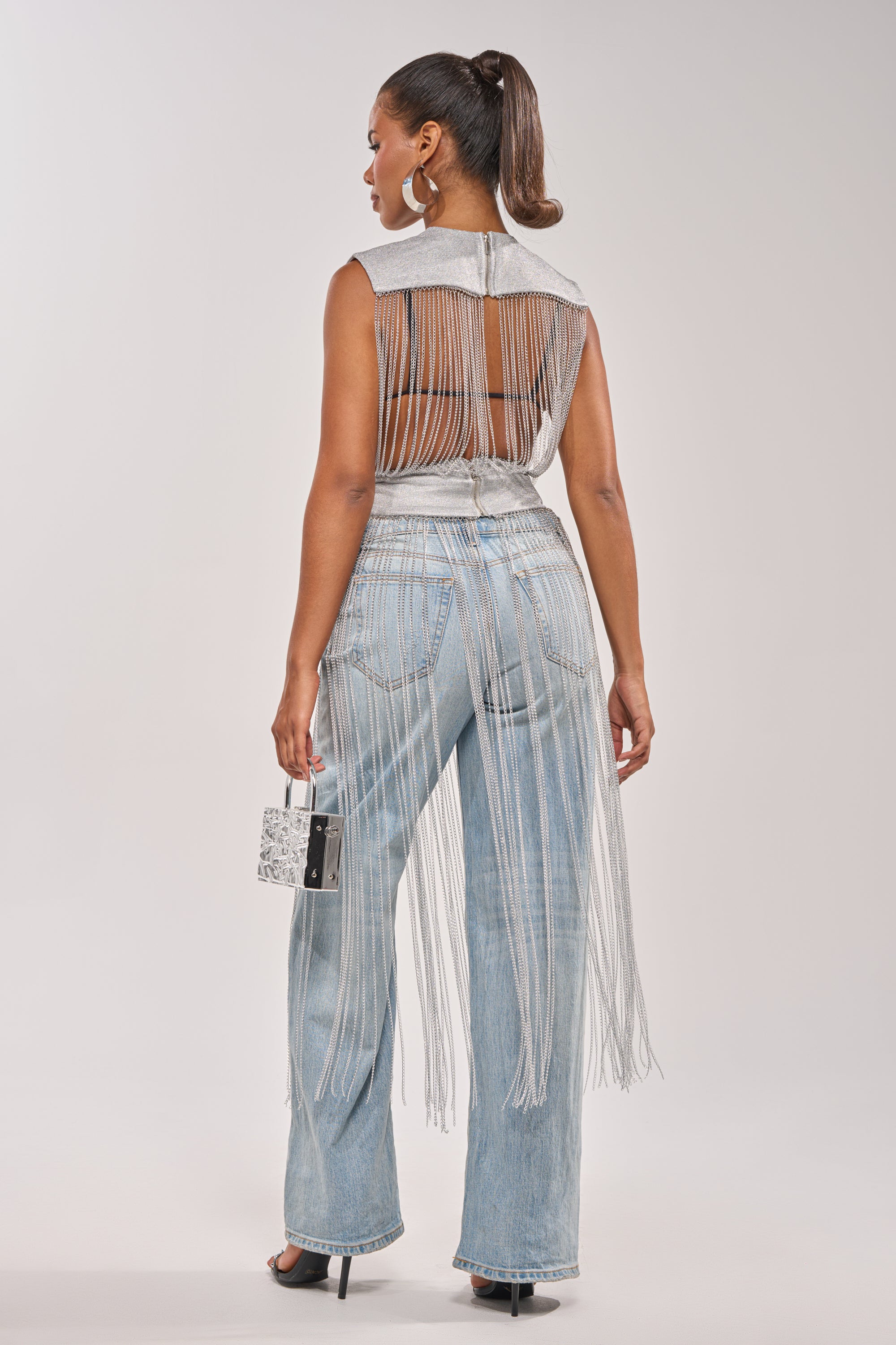 A woman faces away in the ARK SLEEVELESS CHAIN TOP—a bold festival statement—styled with light blue wide-leg jeans, black heels, hoop earrings, a metallic handbag, and her hair in a high ponytail. 6