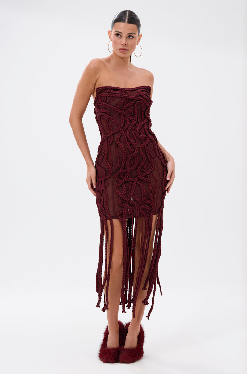 A woman models the ALL OVER THE PLACE BRAIDED MINI DRESS in burgundy, featuring a strapless design, intricate braided detailing, and long fringe at the hem. She pairs it with large hoops and fluffy burgundy shoes on a plain white backdrop.
