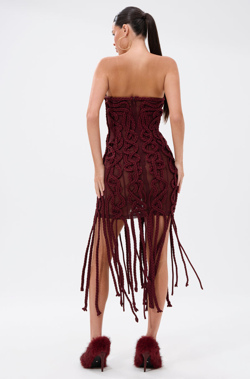 A woman with a high ponytail wears the ALL OVER THE PLACE BRAIDED MINI DRESS in dark red, featuring textured patterns and long fringe details. She pairs it with matching fluffy high heels, standing against a plain white background.