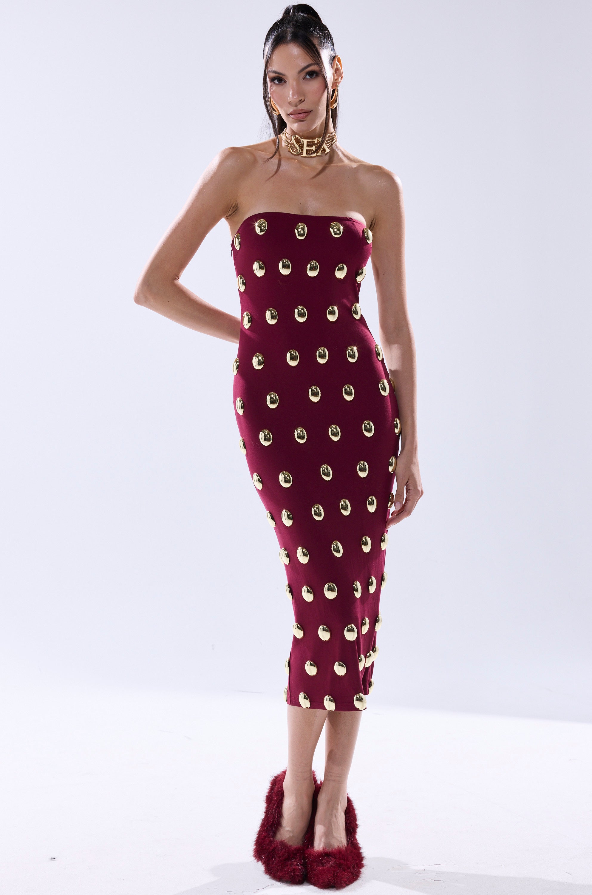 A woman poses in the TIME AFTER TIME MIDI DRESS WITH METAL DETAILING IN BURGUNDY, a strapless fitted piece. She pairs it with red furry shoes and a chunky gold choker, her hair pulled back, against a plain white background.