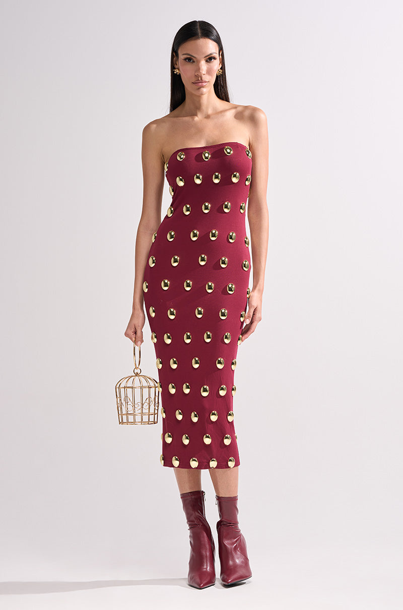A woman stands against a plain light background, wearing the TIME AFTER TIME MIDI DRESS WITH METAL DETAILING IN BURGUNDY, paired with maroon ankle boots and holding a small gold cage-shaped handbag.