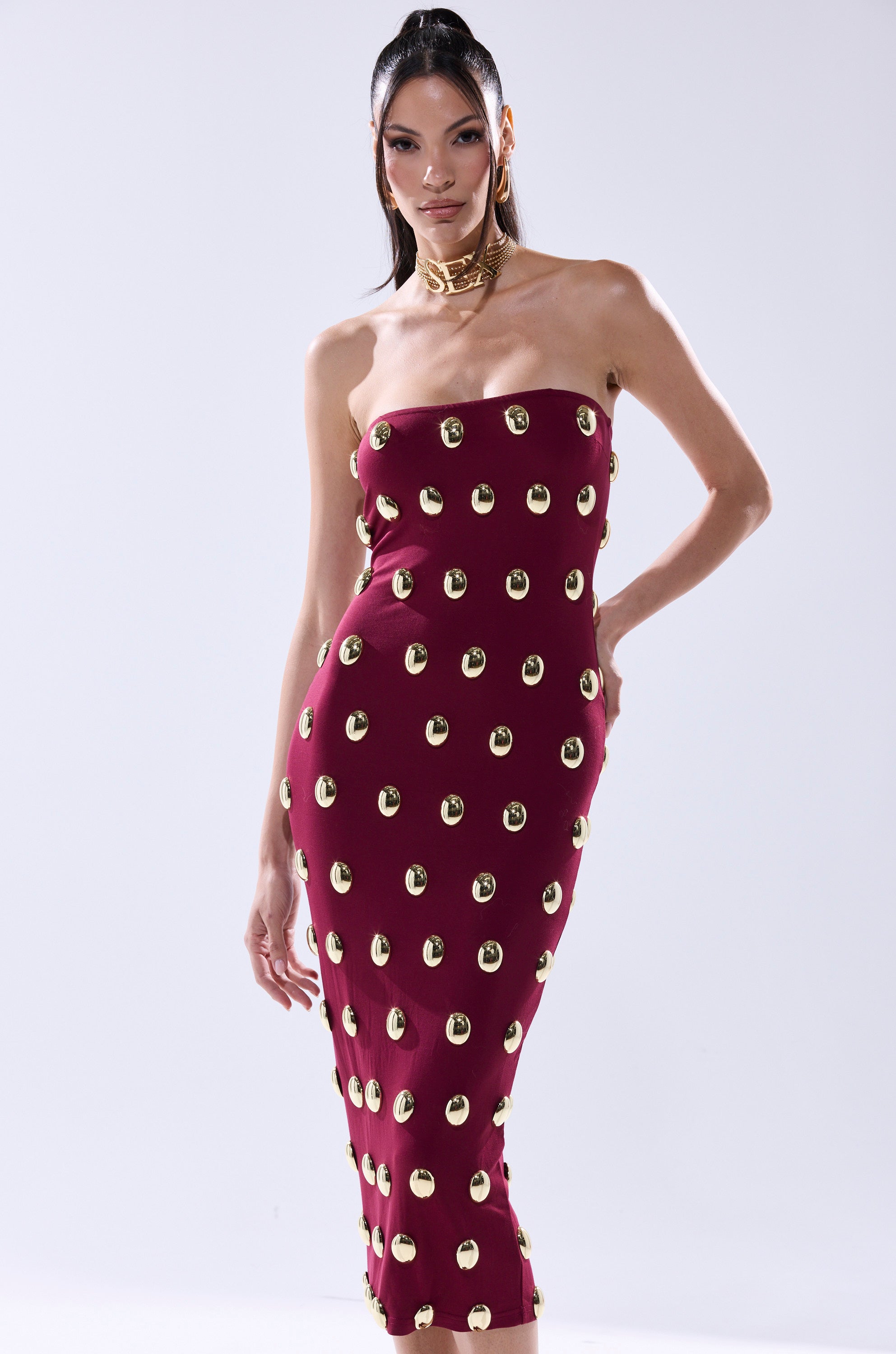 Model showcases the TIME AFTER TIME MIDI DRESS WITH METAL DETAILING IN BURGUNDY—a strapless, form-fitting midi dress featuring evenly spaced gold metal accents—against a plain light background.