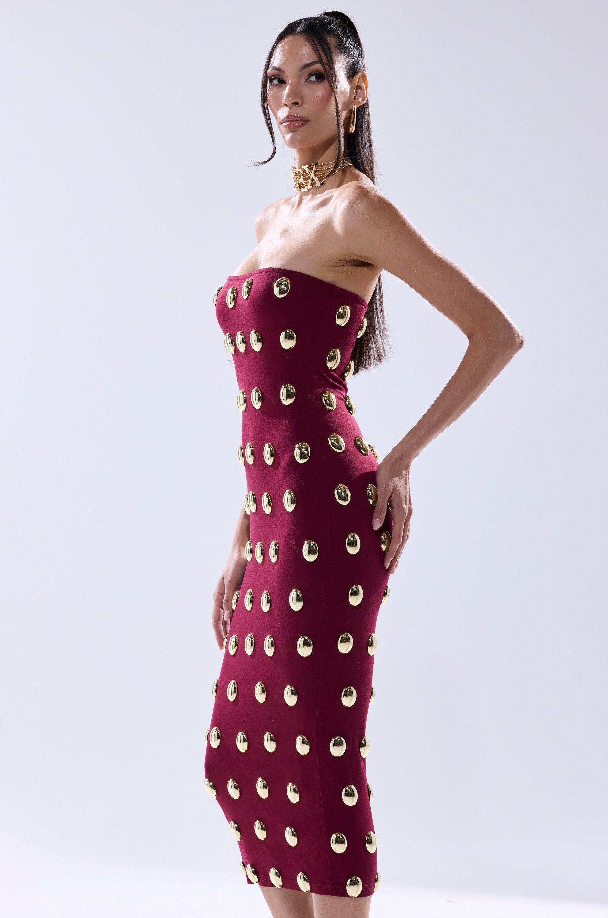 A woman models the TIME AFTER TIME MIDI DRESS WITH METAL DETAILING IN BURGUNDY—strapless, with gold circular accents—paired with a gold choker and pulled-back dark hair, set against a plain light background.