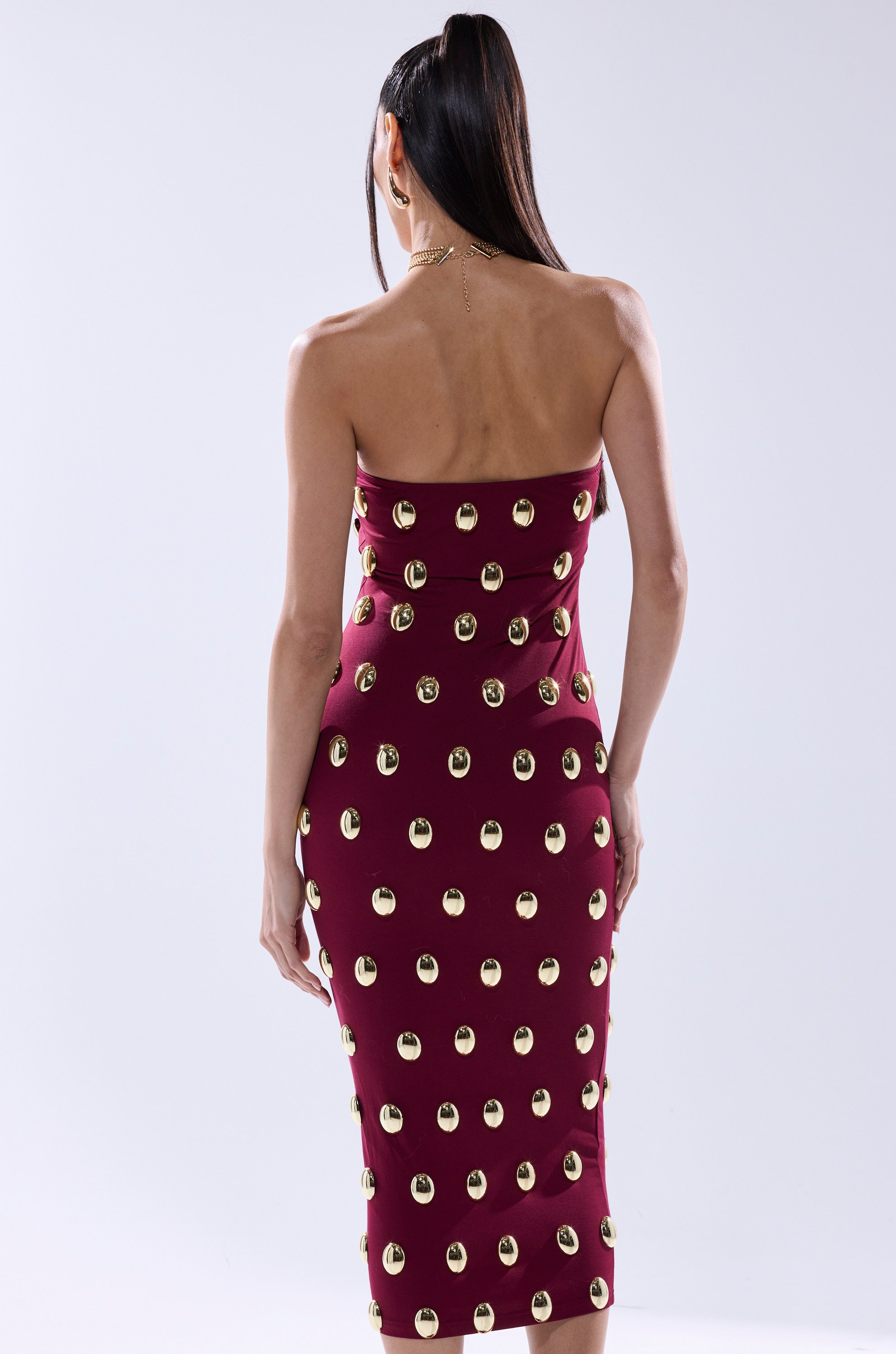 A woman with long dark hair in a high ponytail is seen from the back against a plain light background, wearing the TIME AFTER TIME MIDI DRESS WITH METAL DETAILING IN BURGUNDY, a strapless fitted burgundy midi dress with gold metal accents.