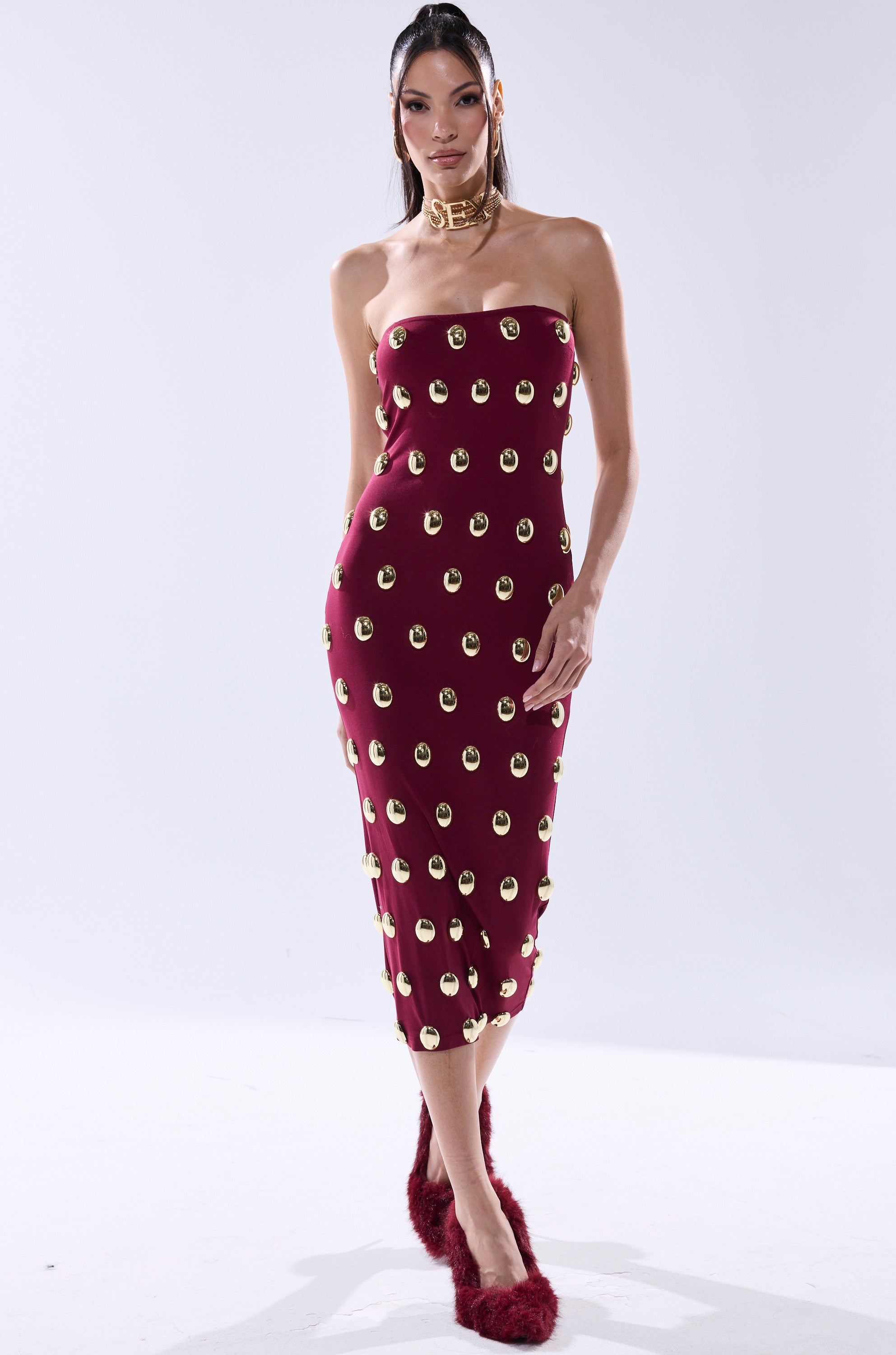 A woman models the TIME AFTER TIME MIDI DRESS WITH METAL DETAILING IN BURGUNDY, styled with a chunky gold choker and maroon fuzzy heels, posing against a plain white background.