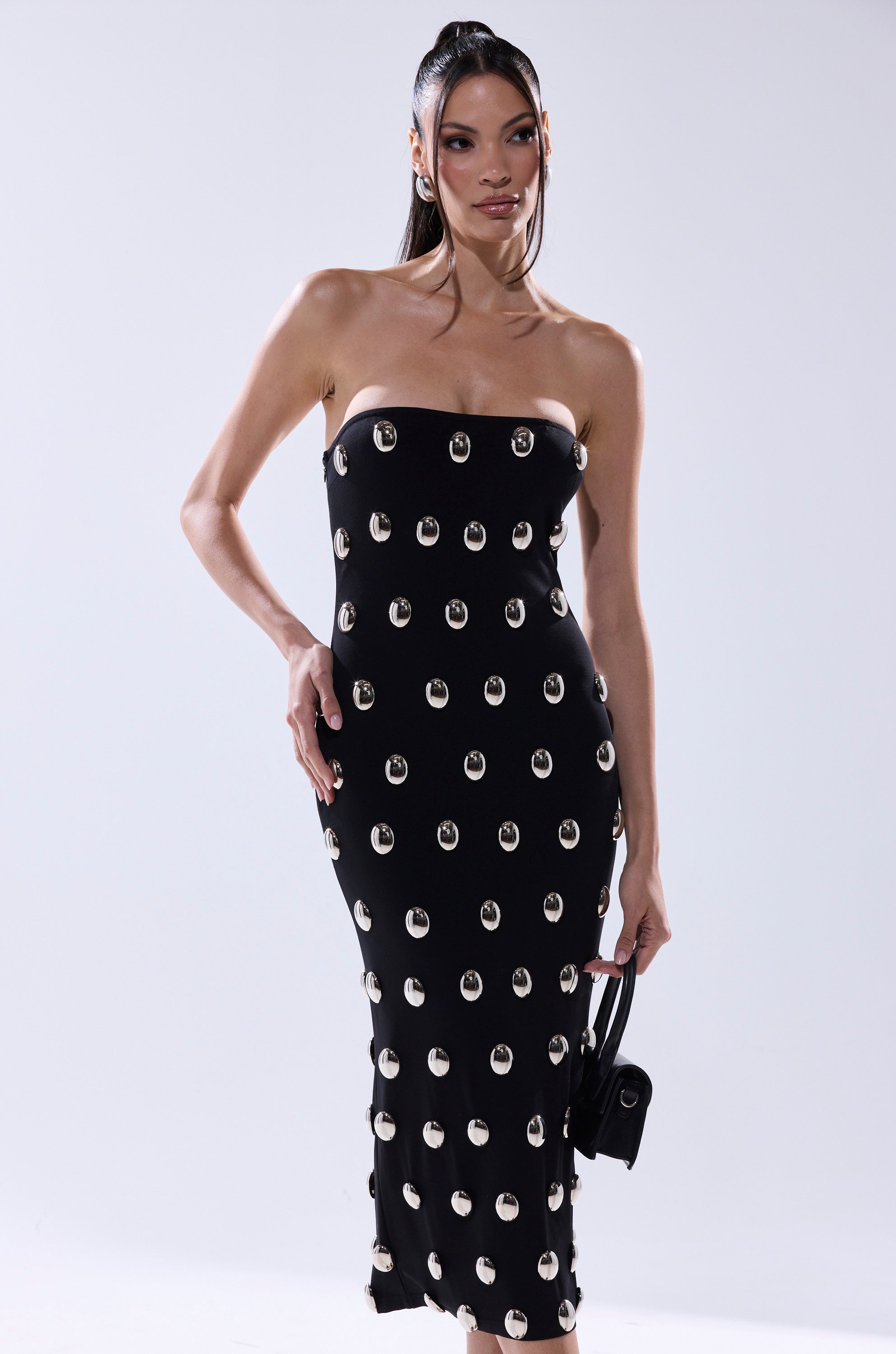 A woman poses against a light background in the TIME AFTER TIME MIDI DRESS WITH METAL DETAILING, a strapless, fitted black bodycon dress with large metallic accents, and holds a small black handbag in her left hand.