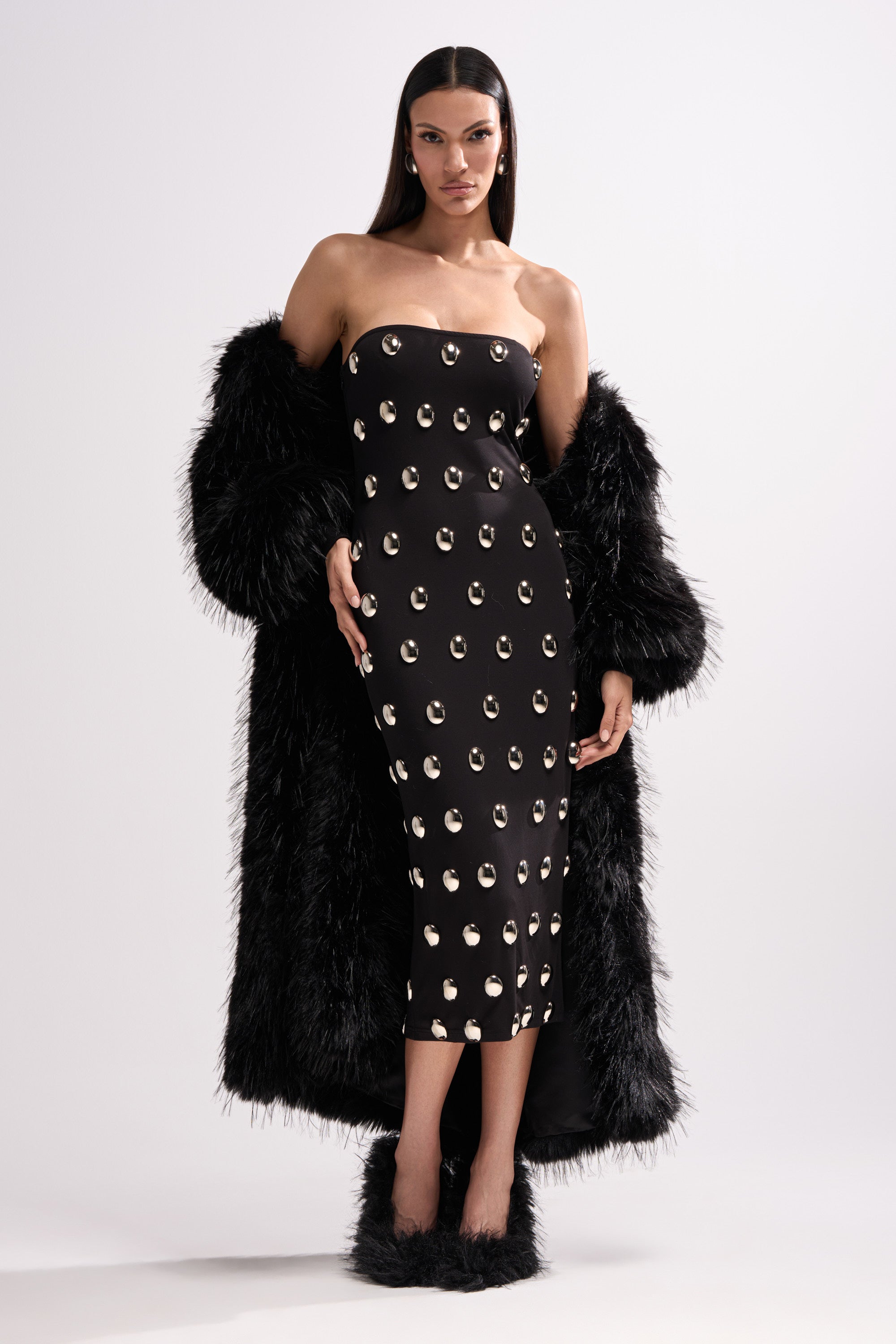 A woman poses against a white background in the strapless TIME AFTER TIME MIDI DRESS WITH METAL DETAILING, styled with a long black faux fur coat over her arms and matching black furry heels.