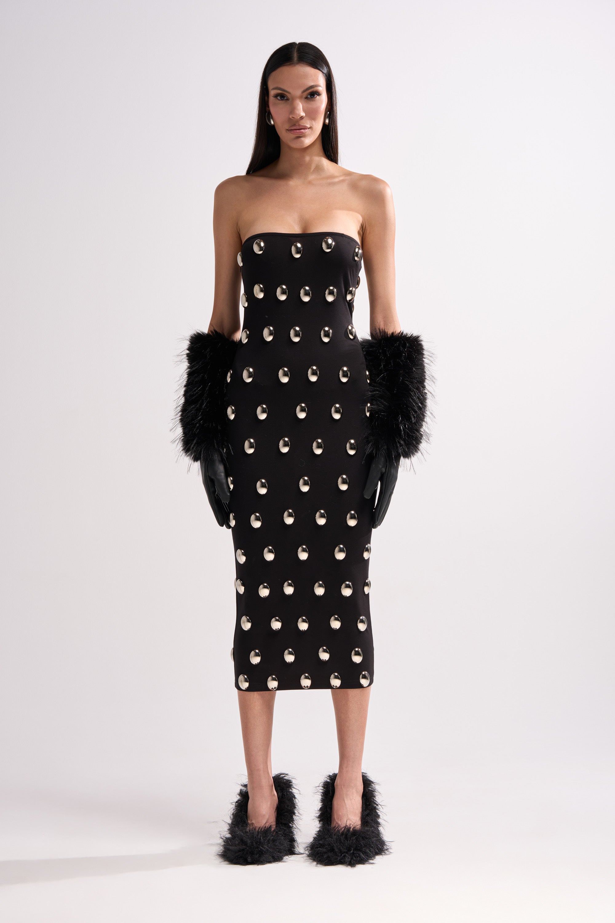 A woman with straight dark hair and a neutral expression stands against a plain background wearing the TIME AFTER TIME MIDI DRESS WITH METAL DETAILING, paired with black fluffy gloves and matching black fluffy shoes.