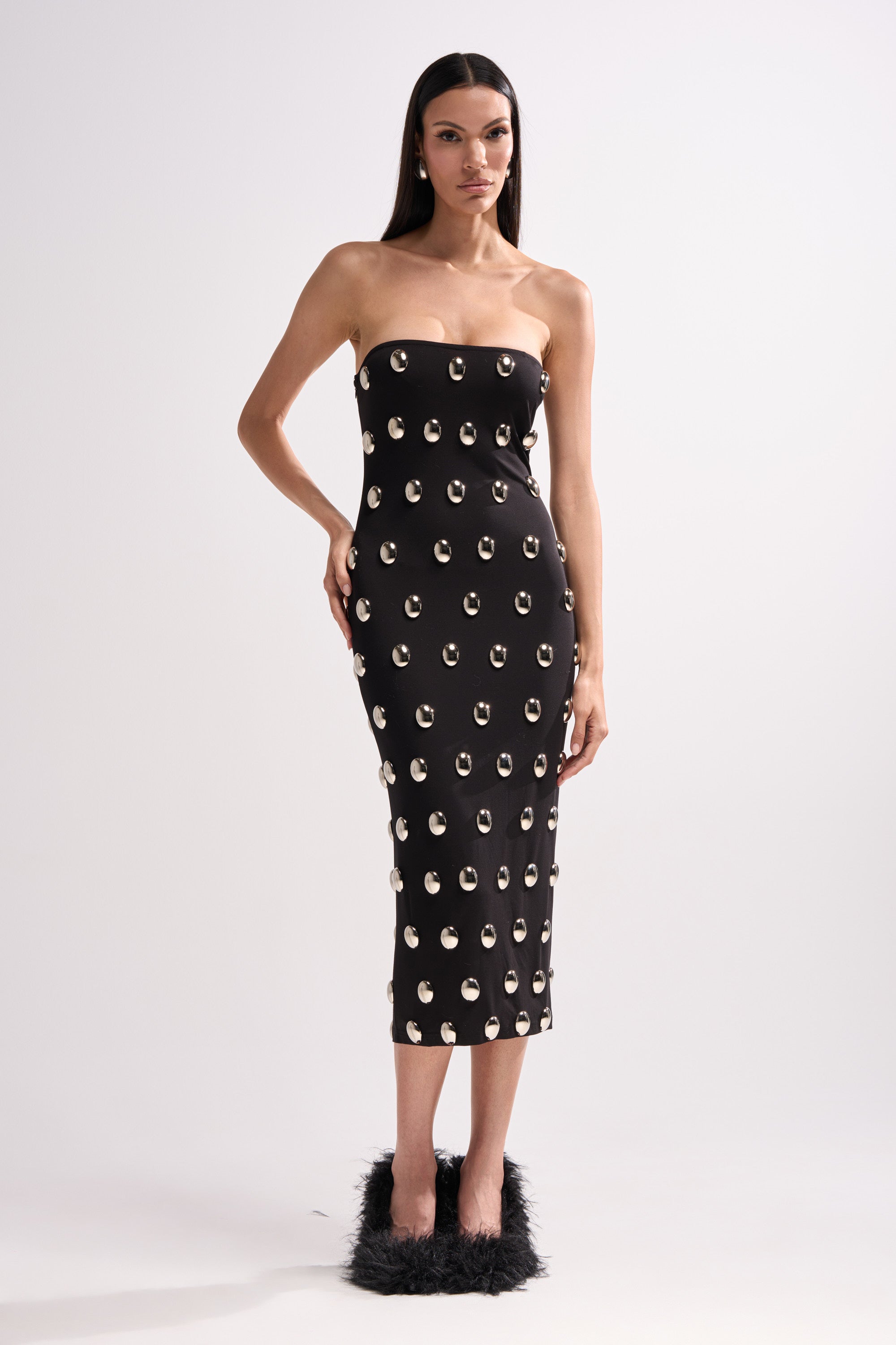 A woman poses against a plain background in the TIME AFTER TIME MIDI DRESS WITH METAL DETAILING, a strapless, form-fitting black bodycon dress with metallic accents and silver embellishments, paired with black fluffy heels.