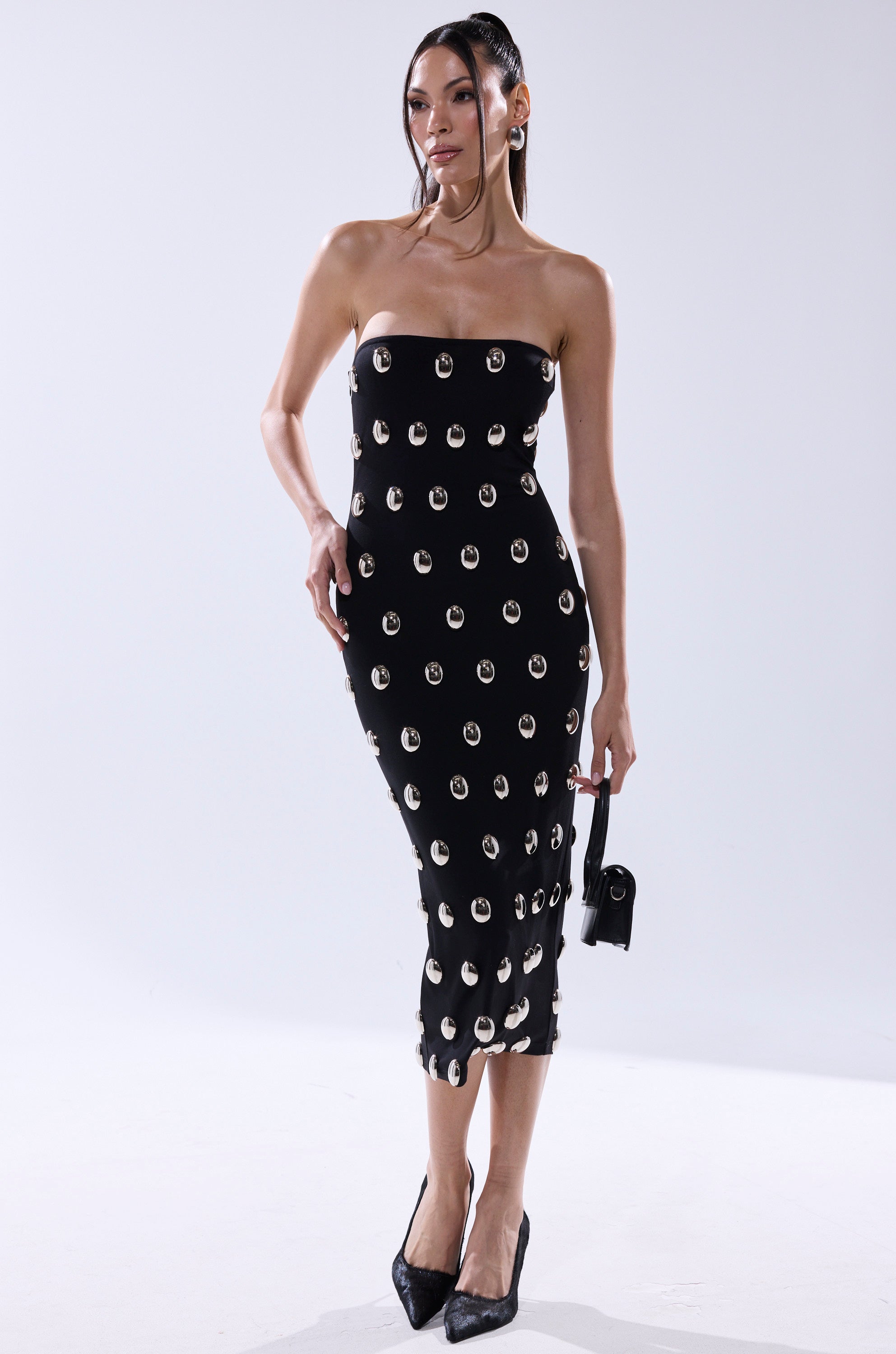 A woman poses against a plain white background wearing the TIME AFTER TIME MIDI DRESS WITH METAL DETAILING, styled with black pointed heels and a small black handbag.