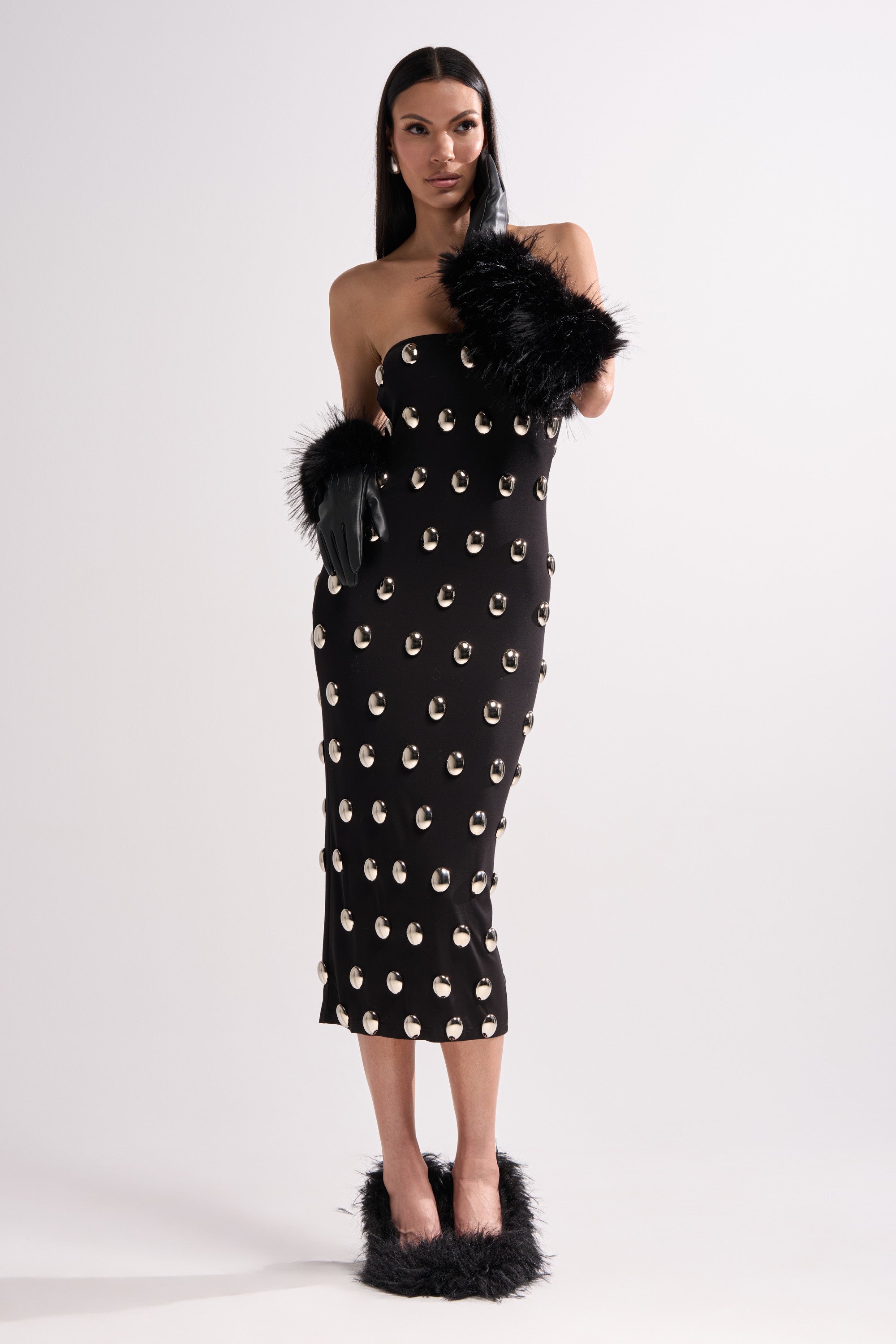 A woman wears the TIME AFTER TIME MIDI DRESS WITH METAL DETAILING, a strapless black bodycon midi dress featuring metallic and circular silver accents, paired with black gloves with feathered cuffs and black feathered shoes, against a white background.