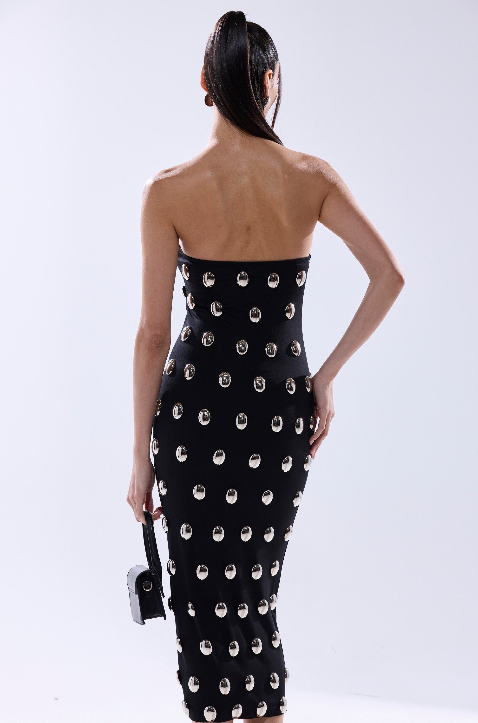 A woman faces away, wearing the TIME AFTER TIME MIDI DRESS WITH METAL DETAILING, a strapless black bodycon style. She holds a small black handbag, her long dark hair in a high ponytail, set against a plain light background.