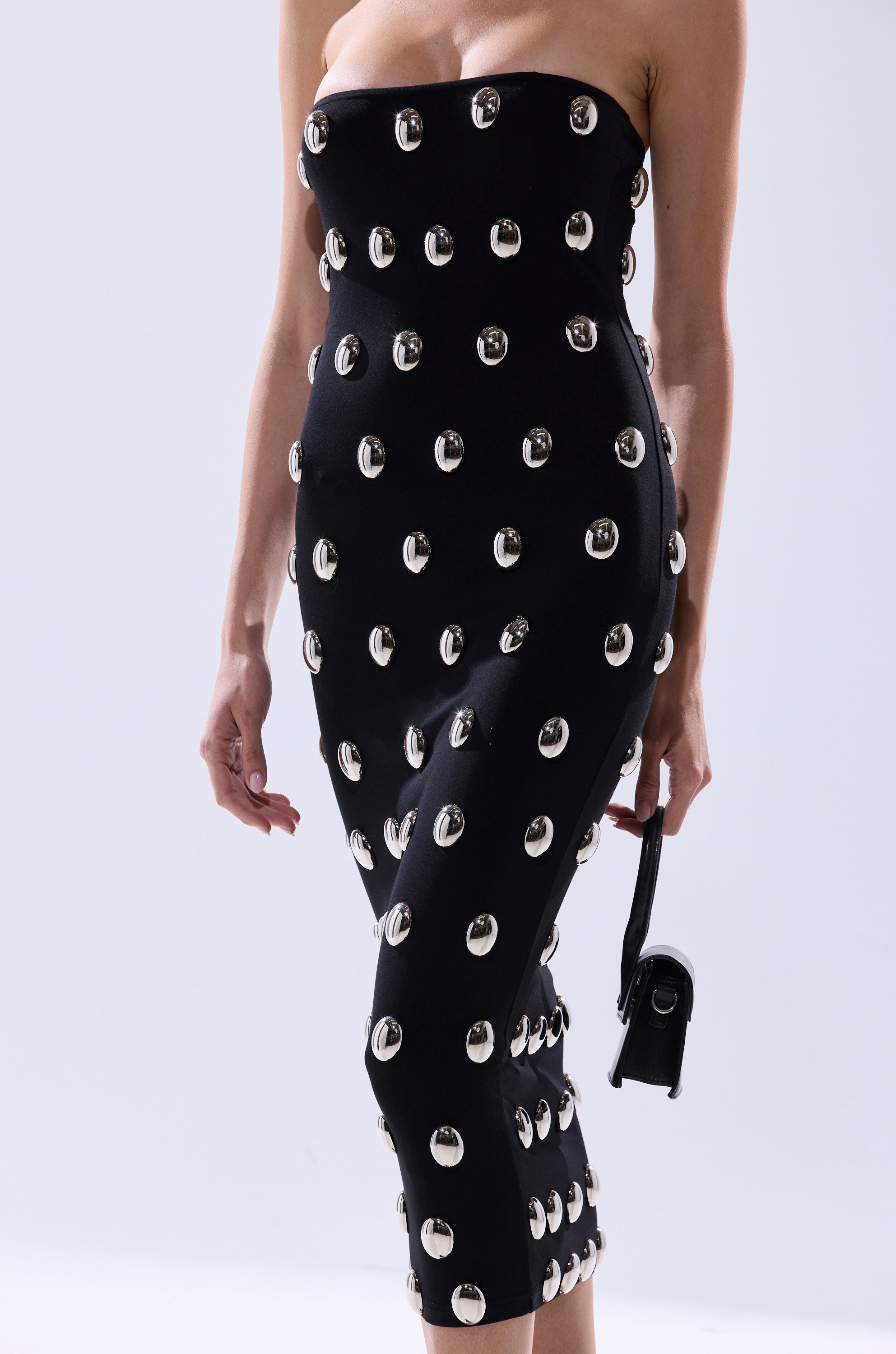 A person wears the TIME AFTER TIME MIDI DRESS WITH METAL DETAILING, a strapless fitted black dress embellished with large silver grommets, and holds a small black handbag against a plain light background.