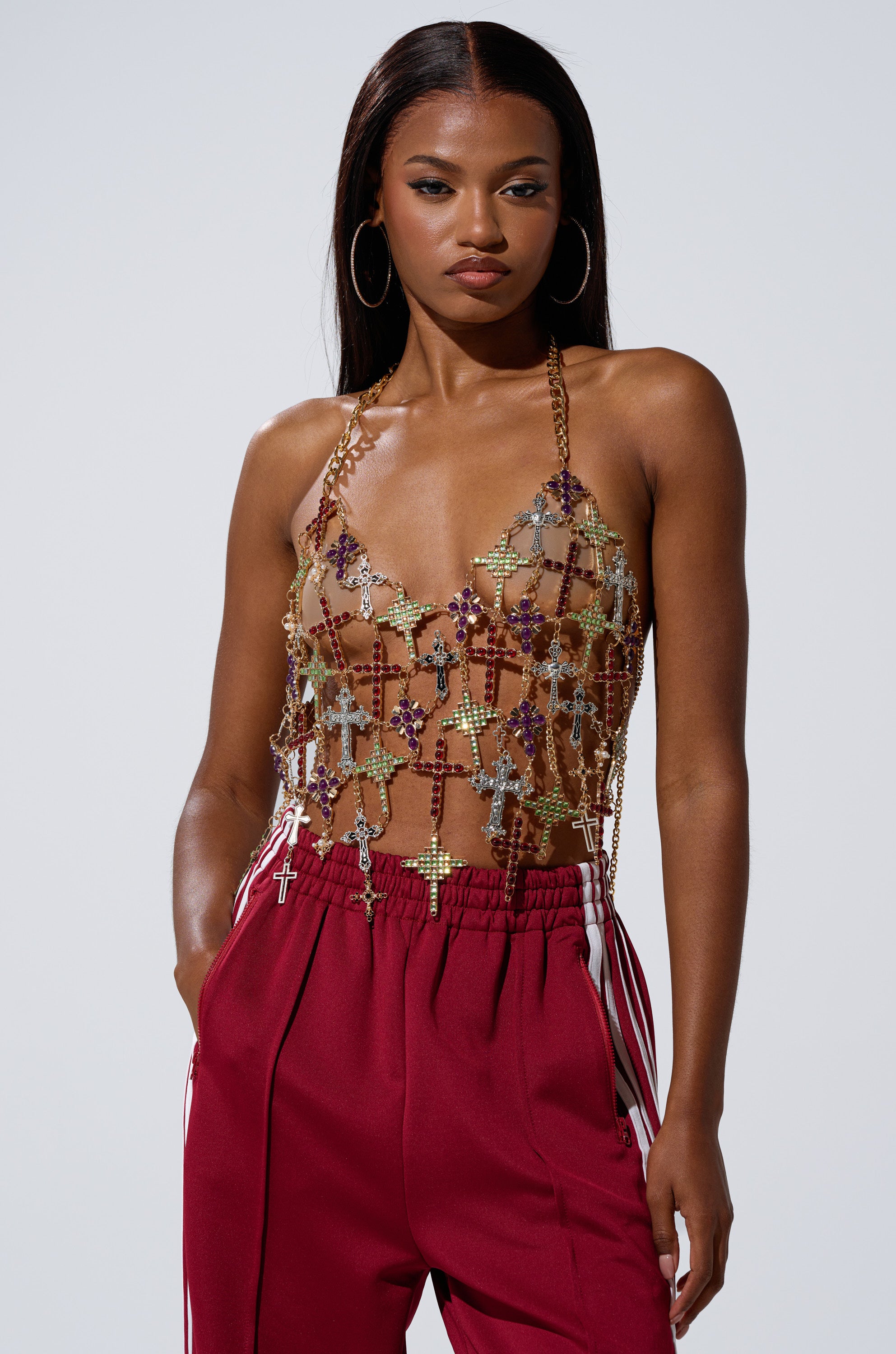 A woman with long dark hair models the BLESSED BRALETTE, a jeweled sheer halter top with cross patterns, paired with red drawstring pants featuring white stripes, posing against a plain light background.