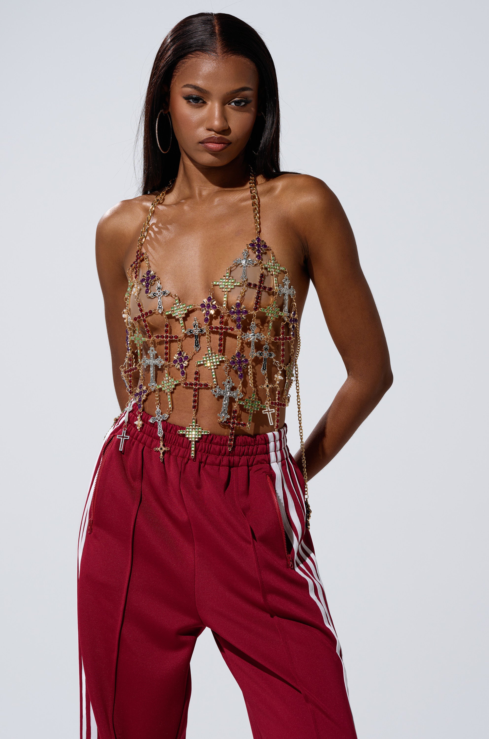 A woman exudes confidence in the BLESSED BRALETTE, a gold chain top with sheer beaded details, paired with high-waisted maroon pants with white side stripes. She accessorizes with large hoops and poses against a light gray background.